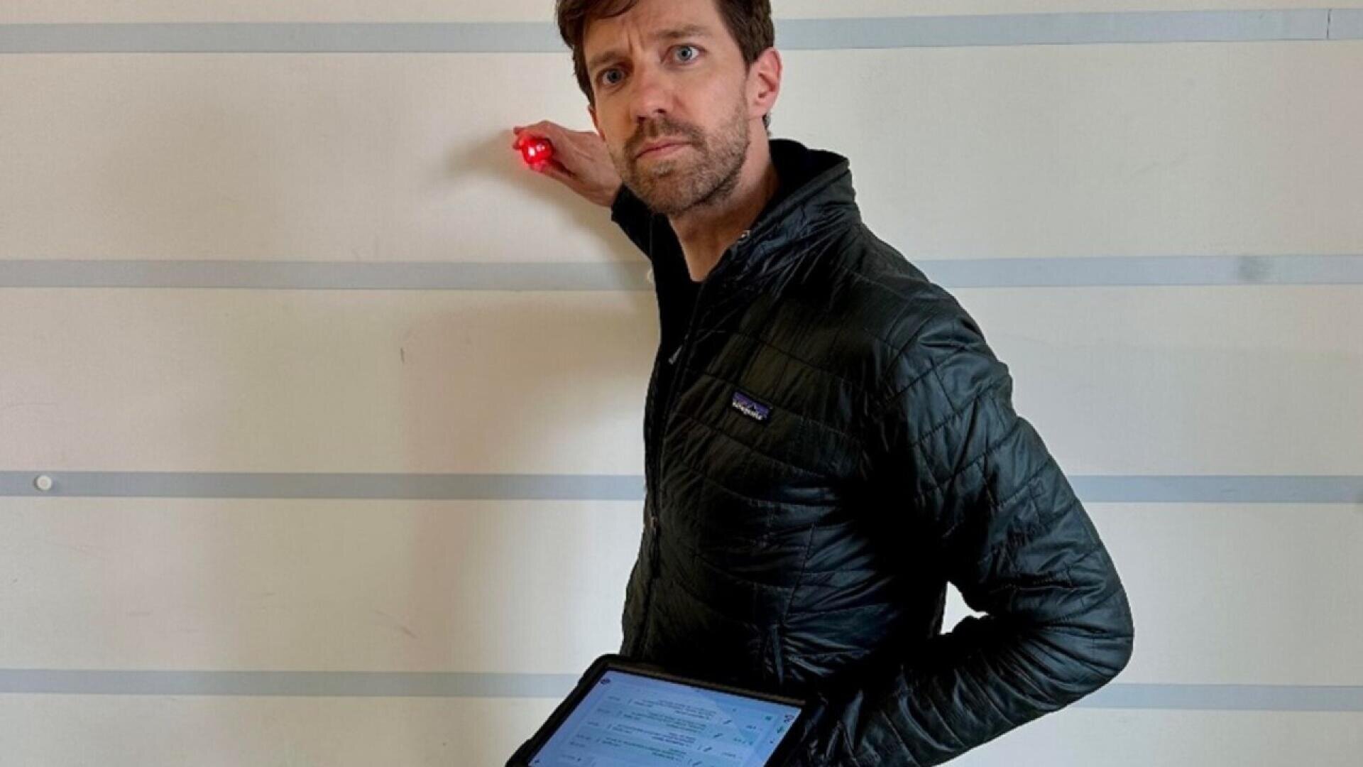 A man in a black jacket holds a tablet in one hand and uses a red light device against a white wall with horizontal lines, looking back over his shoulder.