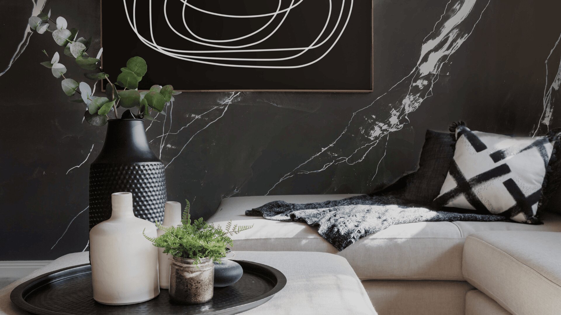 Modern living room with a light beige sofa, black and white abstract art, black and white patterned pillows, a cozy gray throw, and decorative vases with greenery on a round black tray. Marble-patterned dark wall in the background.
