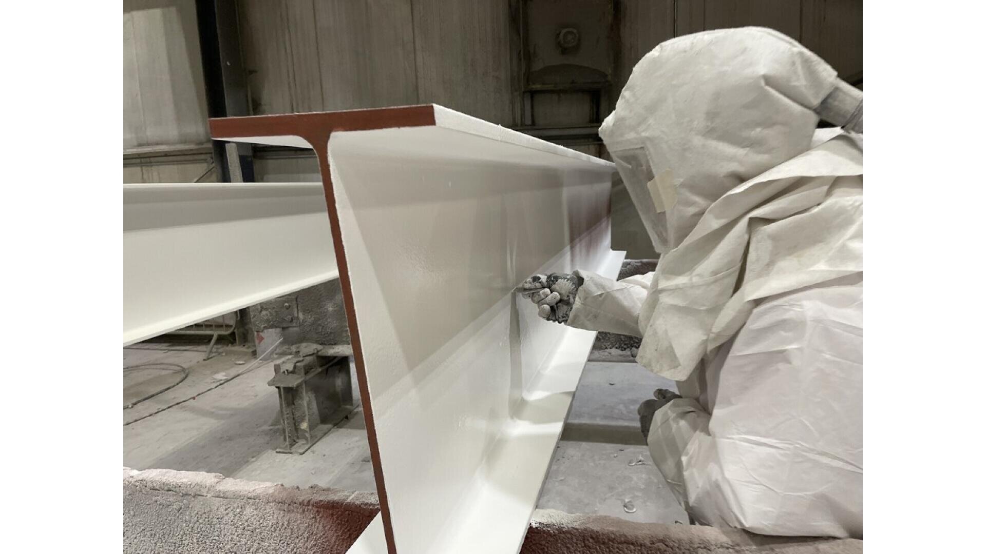 A person in protective gear spray paints a large metal beam white inside an industrial facility. The protective suit covers their entire body, including a hood and face shield.