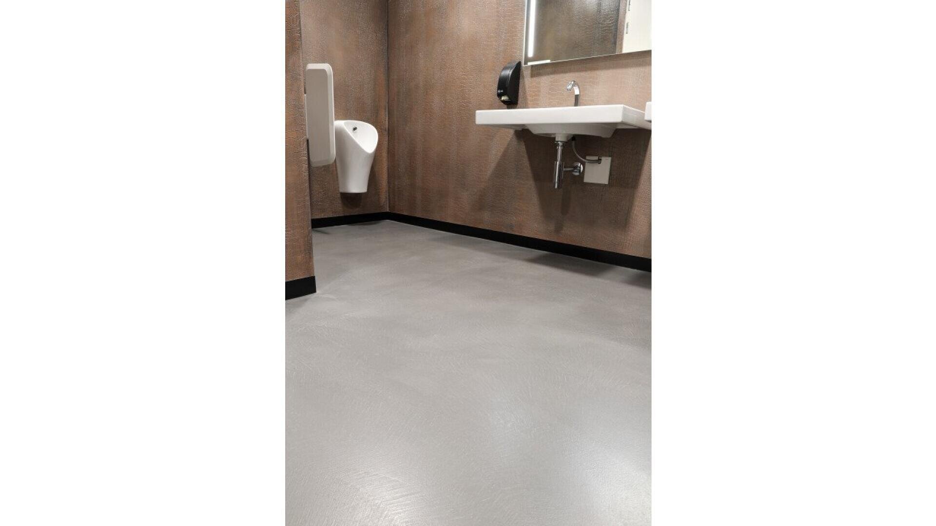 A public restroom with brown textured walls, a light gray polished floor, a white wall-mounted urinal, a white sink with exposed plumbing, a mirror, and a black soap dispenser.