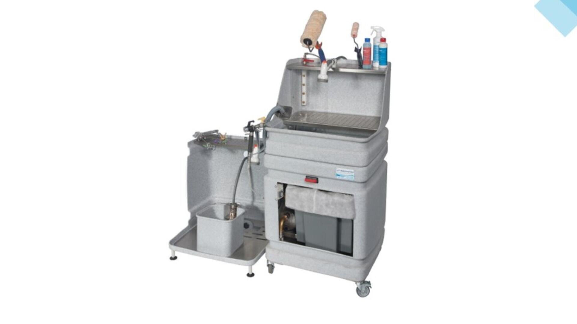 A gray industrial parts washer on wheels with an open lid, cleaning tools, spray bottles, and hoses attached, and a separate side compartment with a drain basin.