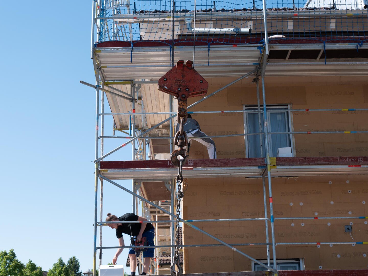 A construction worker stands on scaffolding beside a building under construction, holding onto a large crane hook and chain. Another worker is bent over, working on the lower level. Blue sky in the background.