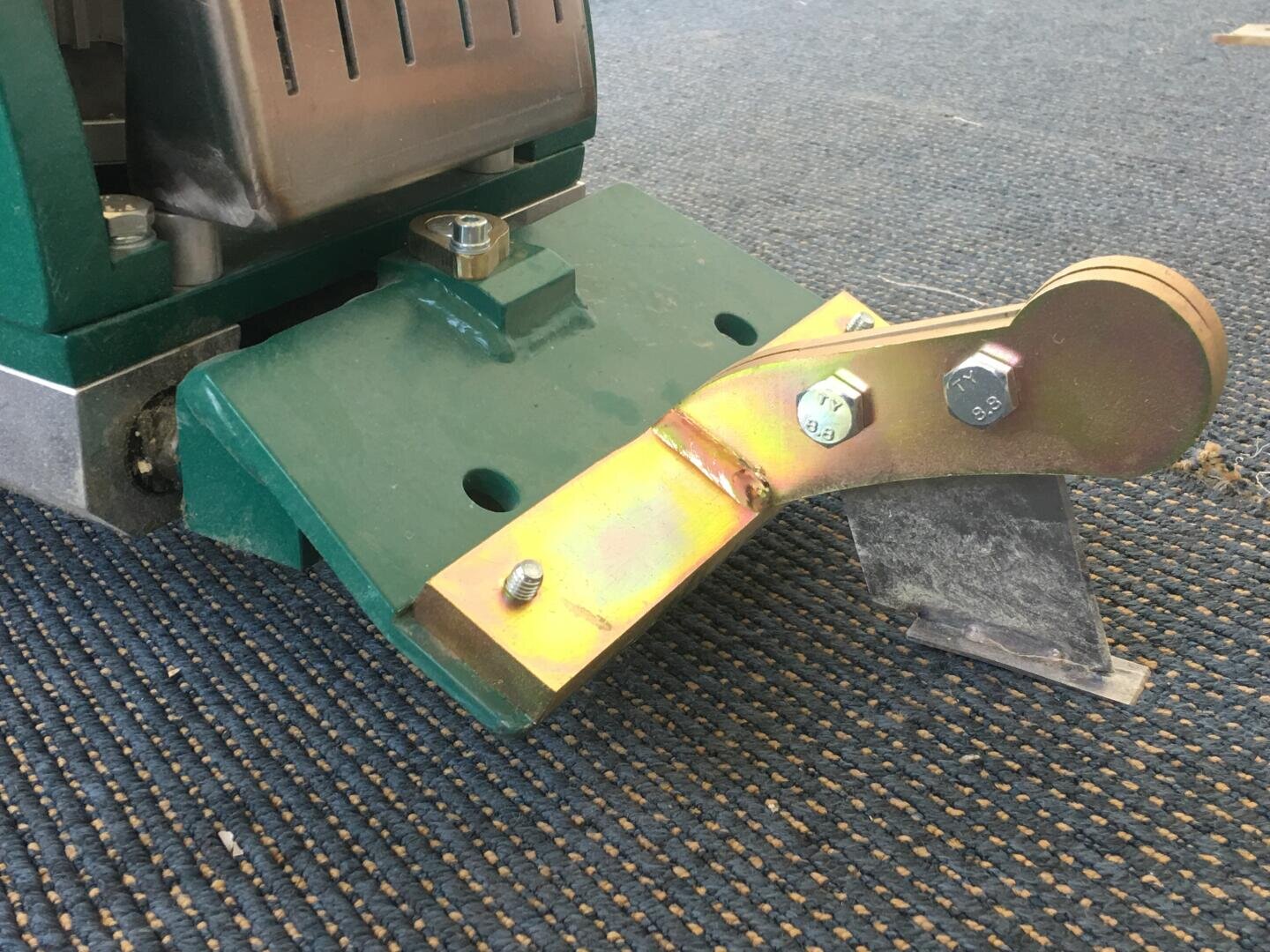 Nahaufnahme eines grün-metallischen Industriemaschinenteils mit Schrauben, montiert auf einer blau strukturierten Oberfläche. Das Teil verfügt über eine abgewinkelte Metallklinge und eine gelbliche Halterung, die mit großen Schrauben befestigt ist.