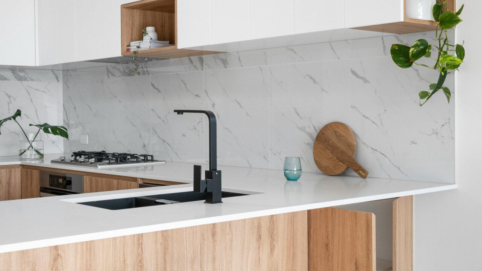 Modern kitchen with light wood cabinets, white countertops, a black faucet, marble backsplash, a round wooden cutting board, glass, and green plants for decoration. The design is clean and minimalistic.