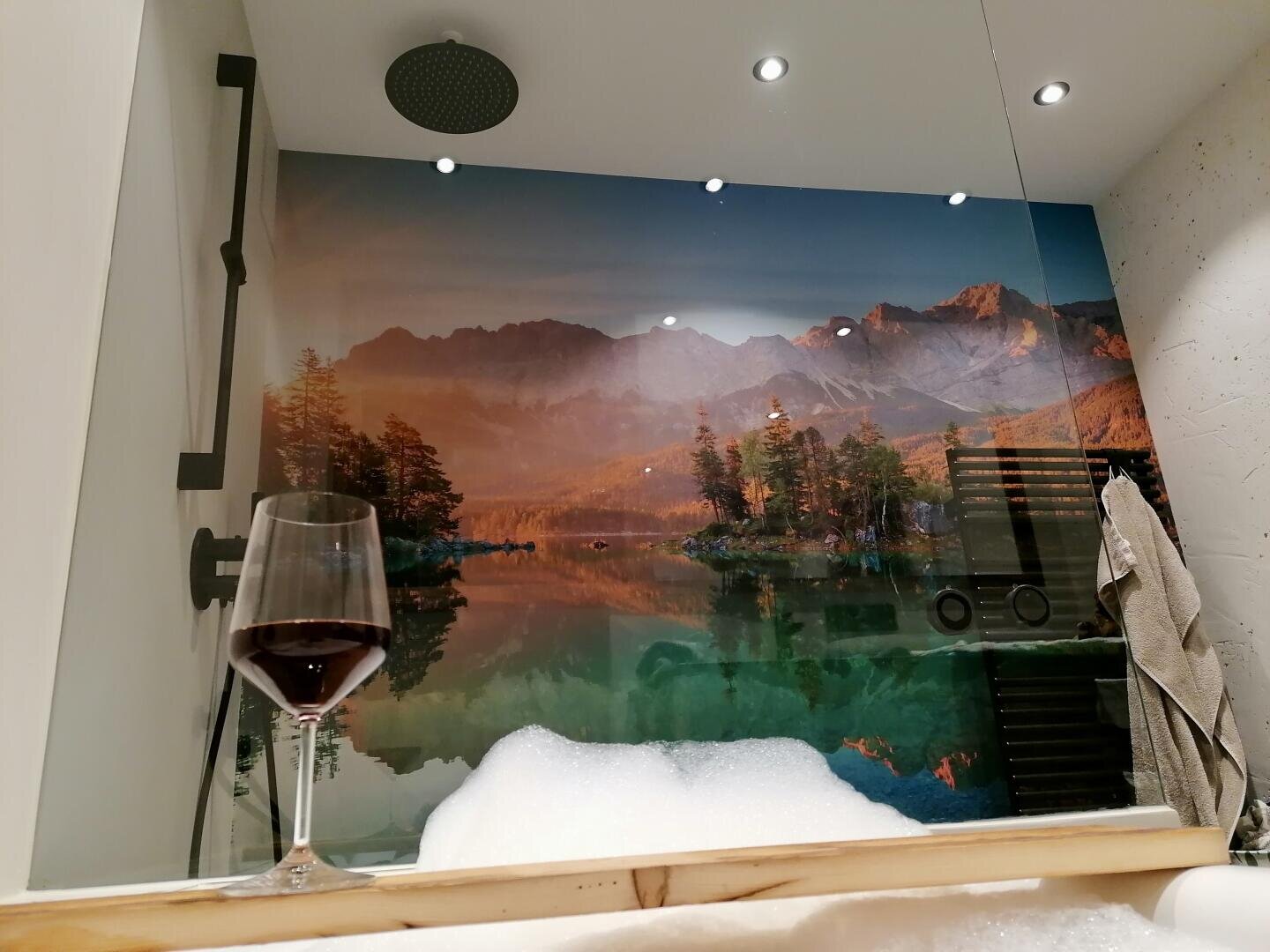 Ein Weinglas steht auf dem Rand einer mit Schaum gefüllten Badewanne, an deren Wand sich ein Wandgemälde einer ruhigen Bergseelandschaft befindet, das eine entspannende Spa-Atmosphäre schafft.