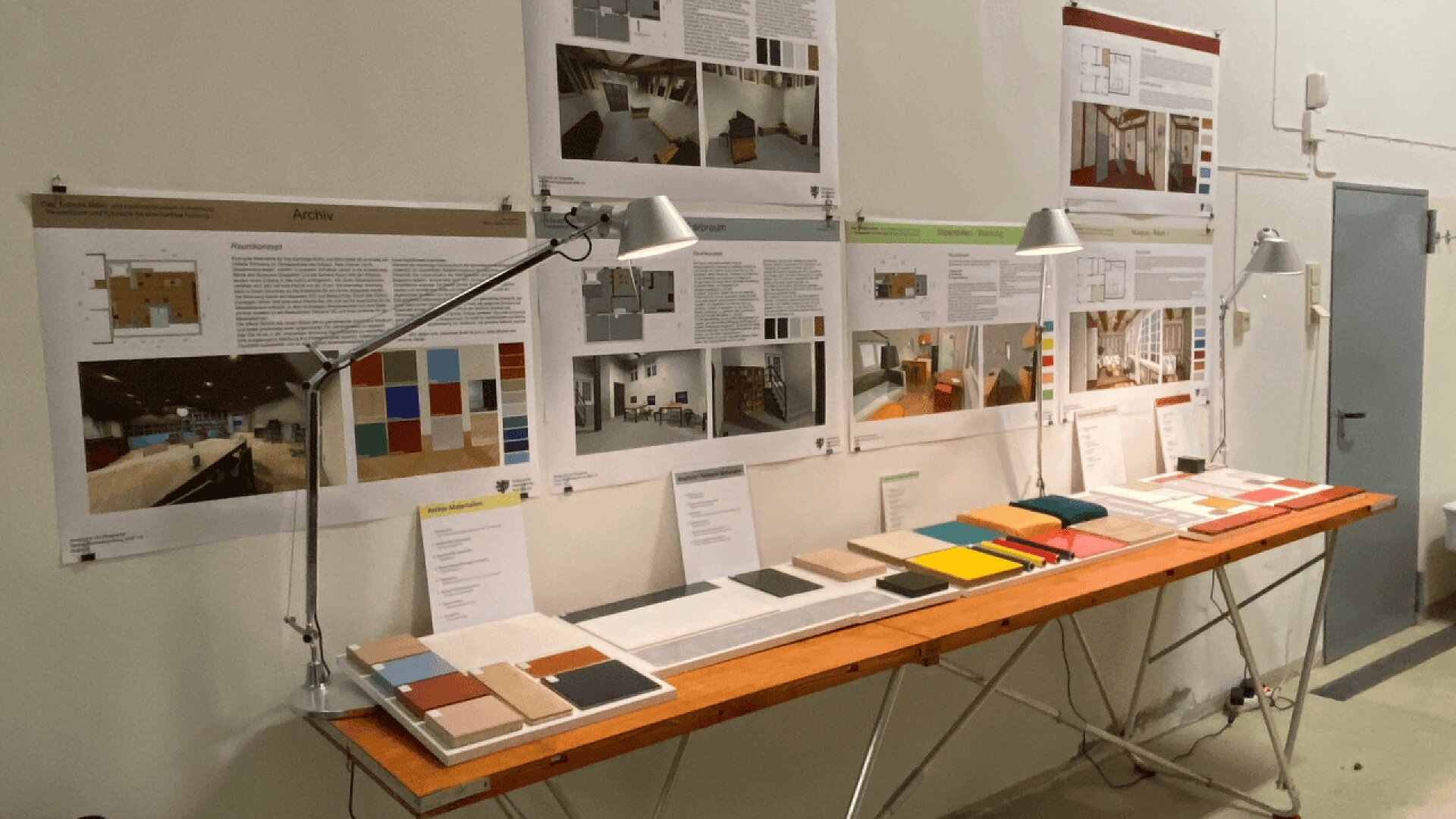 A long table displays color samples and material swatches under desk lamps. Behind it, posters with architectural plans, photos, and color palettes are mounted on a white wall in a design studio environment.