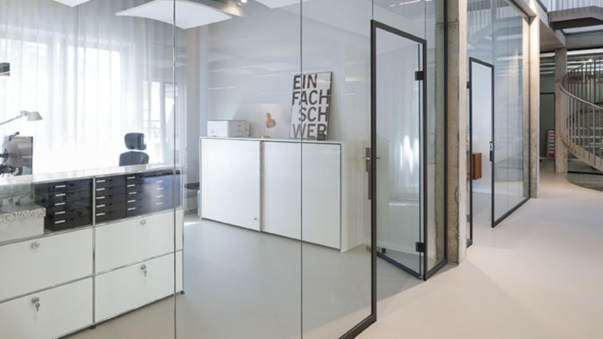 Ein modernes Büro mit Glaswänden, minimalistischen weißen Schränken, einem Schreibtisch mit Stuhl und einem gerahmten Druck, der an einem Schrank lehnt. Der Raum ist hell erleuchtet durch natürliches Licht aus großen Fenstern.