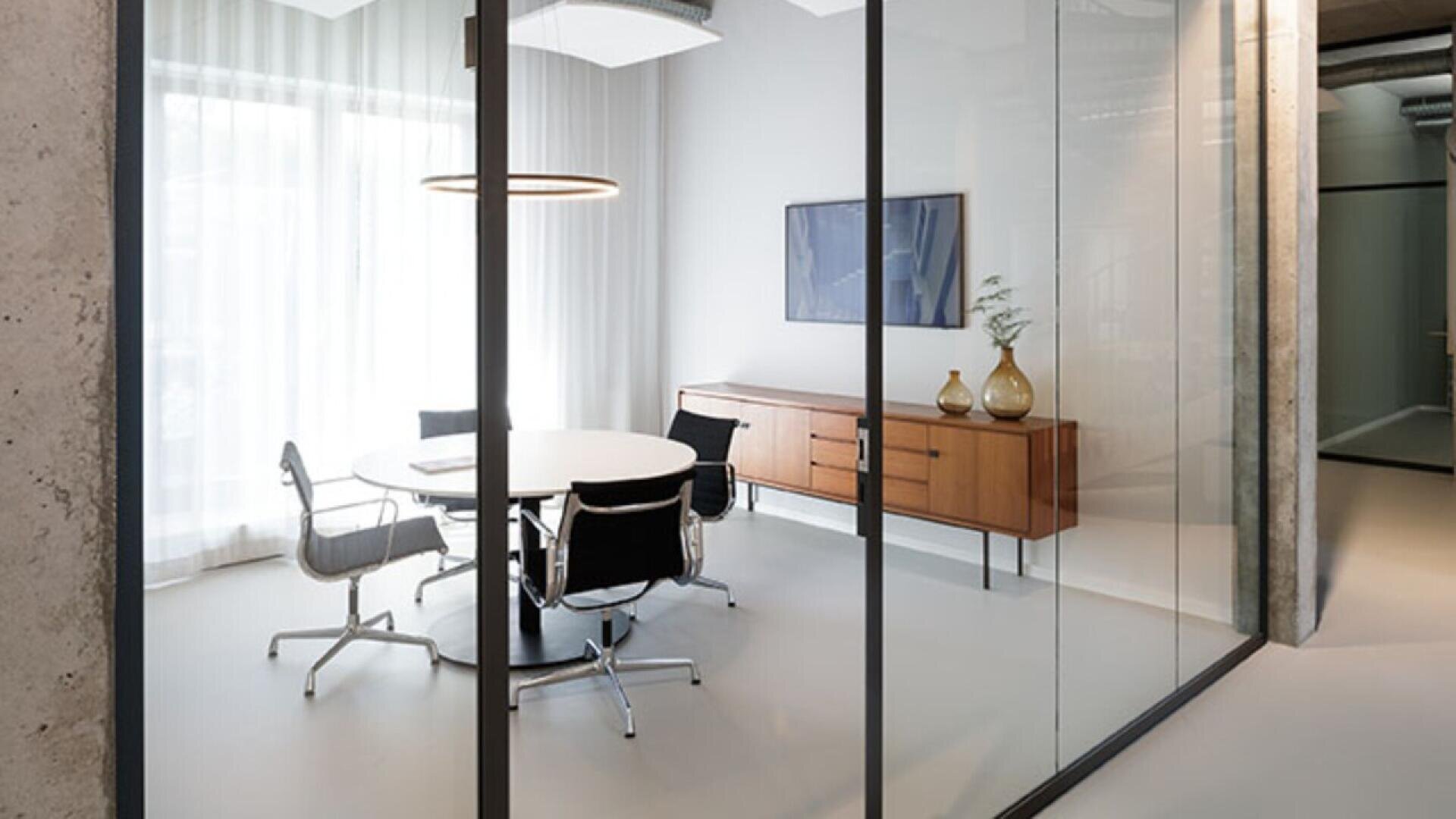 A modern office meeting room with a round white table, four black chairs, a wooden sideboard with decorative vases, a circular ceiling light, and sheer curtains covering a large window.