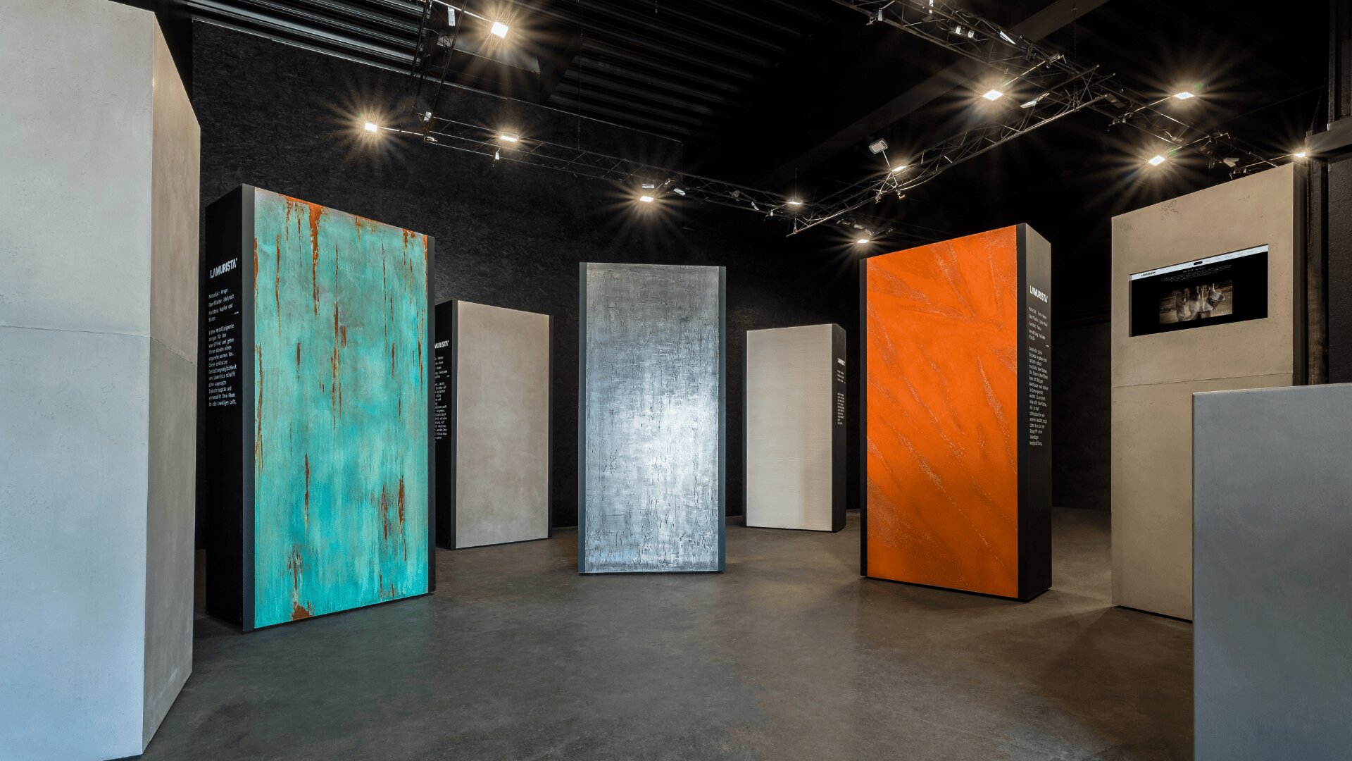 Ein moderner Ausstellungsraum mit hohen rechteckigen Säulen und unterschiedlich strukturierten und farbigen Oberflächen, darunter Türkis, Silber, Orange und Betonoberflächen, die unter Scheinwerfern an einer dunklen Decke ausgestellt sind.