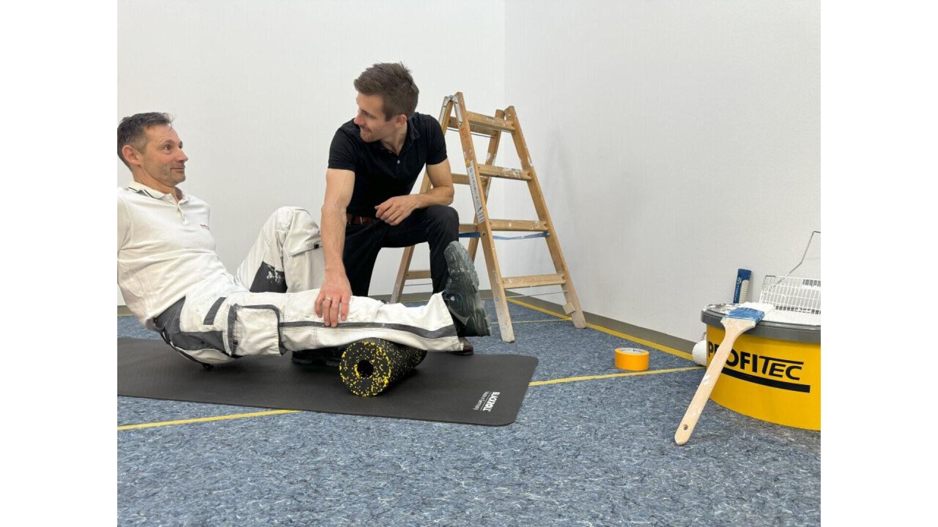 Two men in work clothes are in a room with a ladder and painting supplies. One man sits on a mat using a foam roller under his back, while the other kneels beside him, offering assistance.