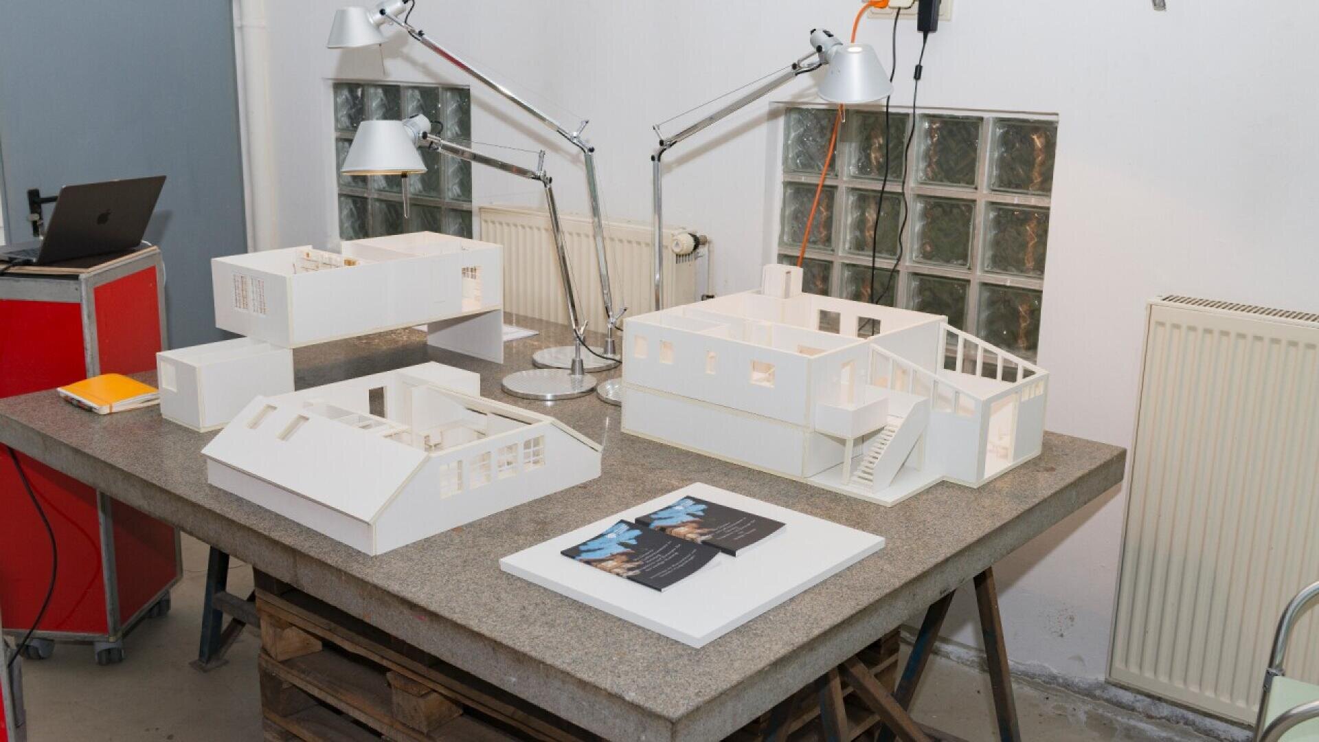 Three white architectural building models are displayed on a concrete table, surrounded by two adjustable desk lamps, magazines, and a notebook in a well-lit workspace with industrial decor.