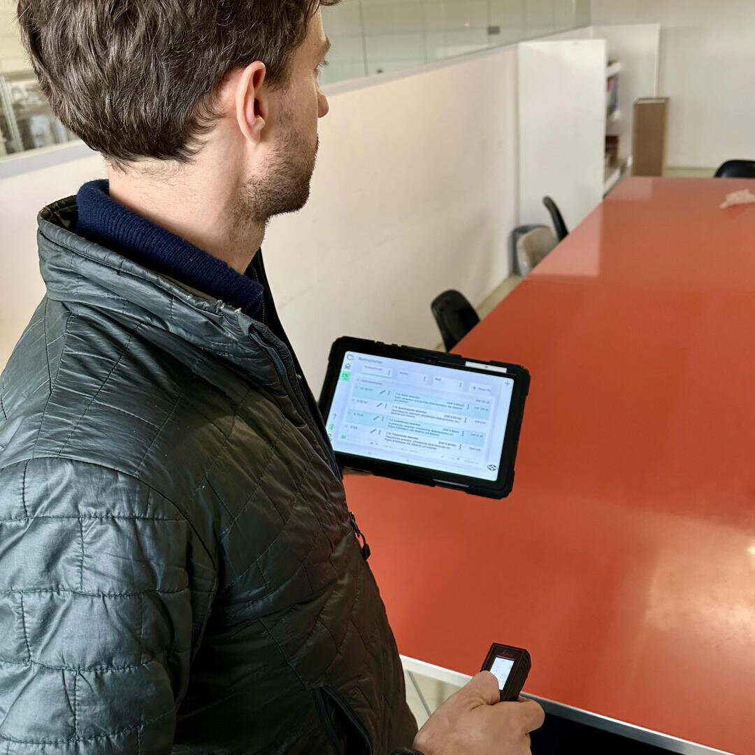 Ein Mann in einer Steppjacke hält ein Tablet mit einem digitalen Formular und einem kleinen Gerät in der Hand, während er neben einem langen roten Konferenztisch in einem Büro steht.