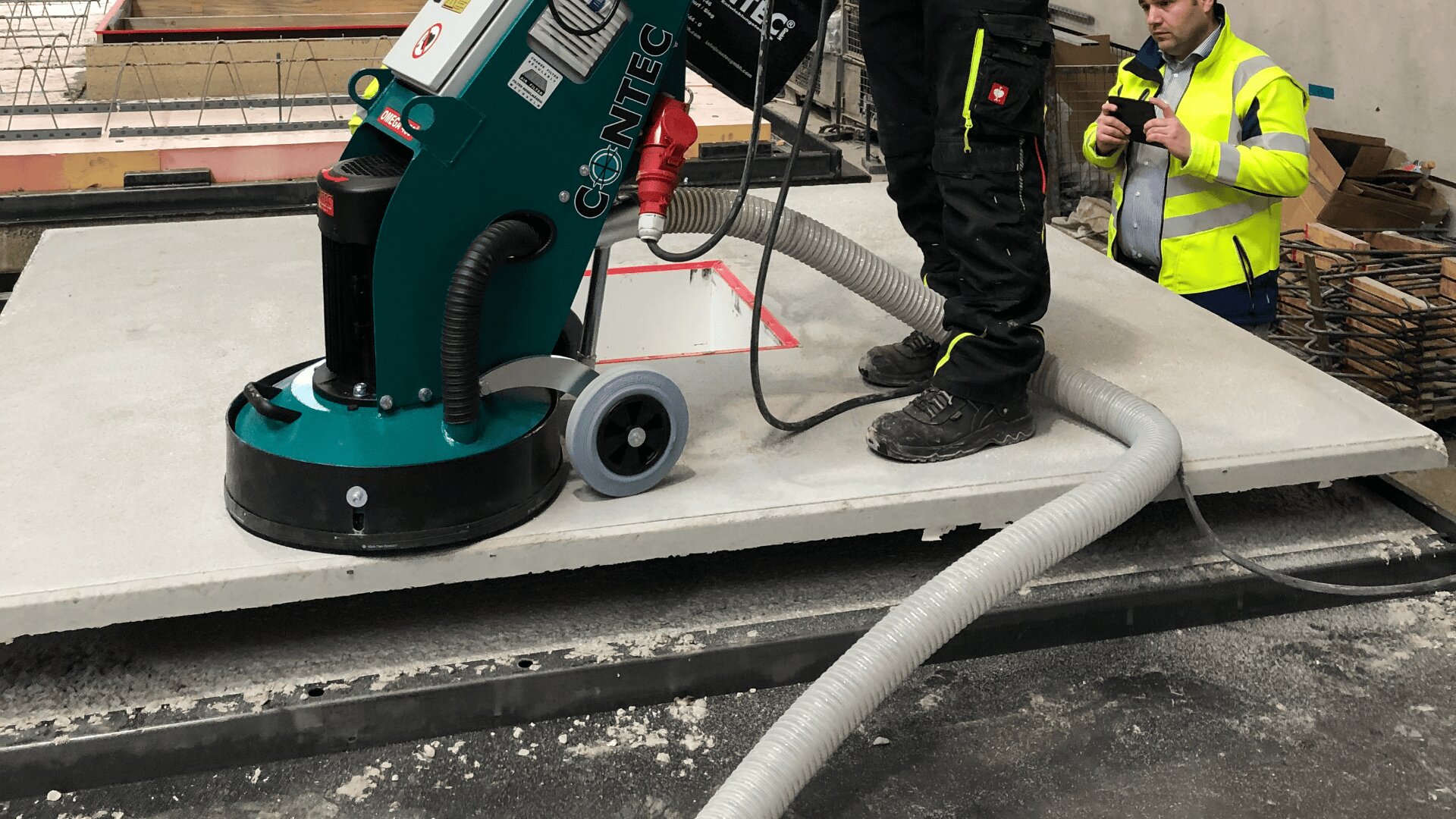 Ein Arbeiter bearbeitet eine Betonplatte mit einer großen grünen Bodenschleifmaschine, während eine andere Person in einer gelben Sicherheitsweste zusieht und sich Notizen macht. An der Maschine sind Schläuche angebracht, um den Staub aufzufangen.