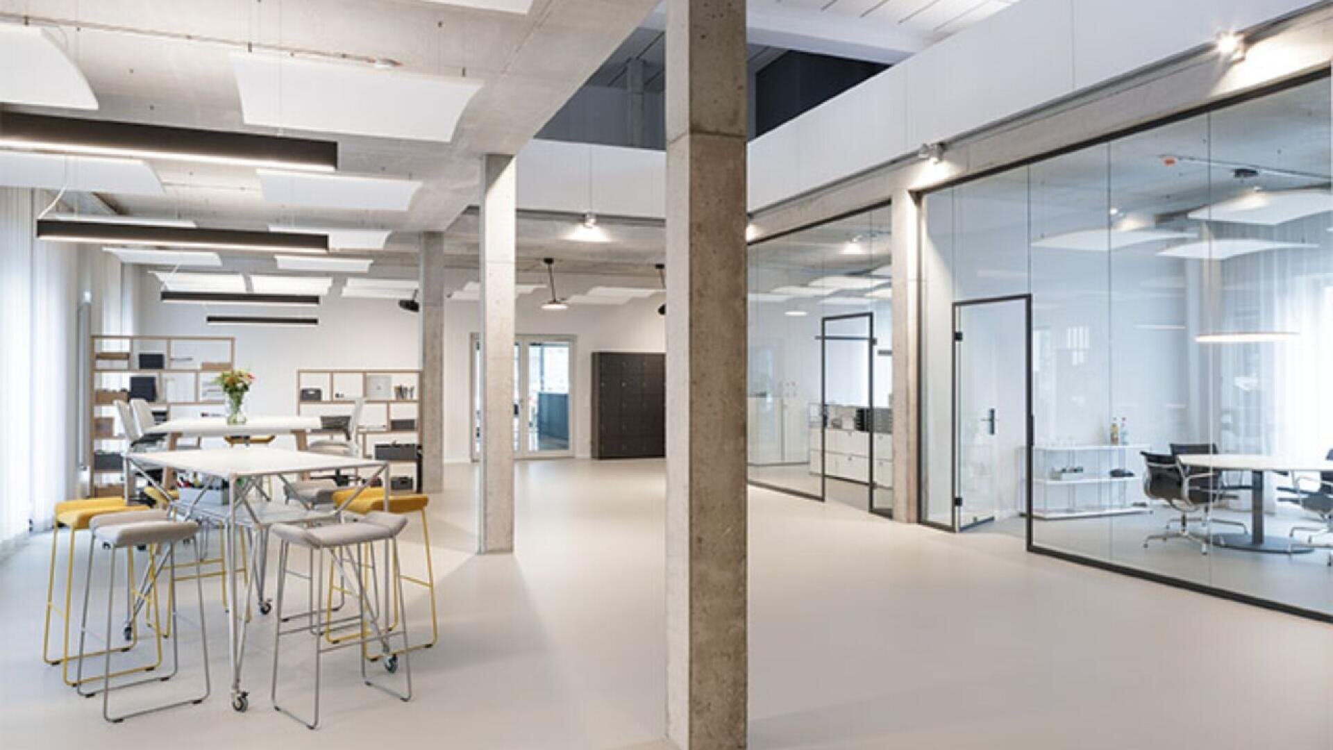 Modern open-plan office with high ceilings, glass-walled meeting rooms, white floors, exposed concrete beams, tall tables with stools, and natural light filling the space. Minimalist and contemporary design.