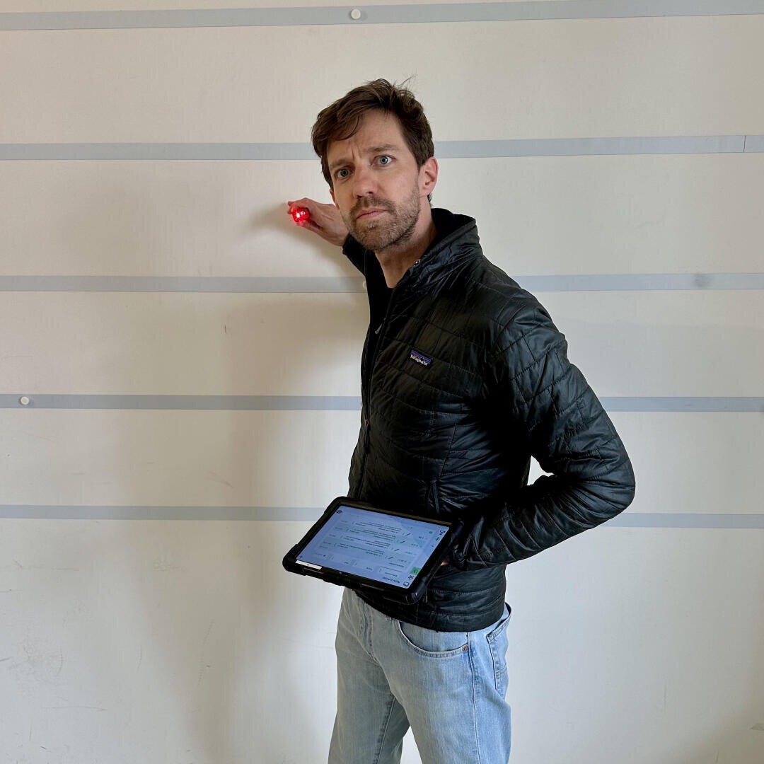 Ein Mann in einer schwarzen Jacke hält in der einen Hand ein Tablet, mit der anderen zielt er mit einem roten Laserpointer auf eine weiß gestreifte Wand und blickt dabei mit ernster Miene in die Kamera.