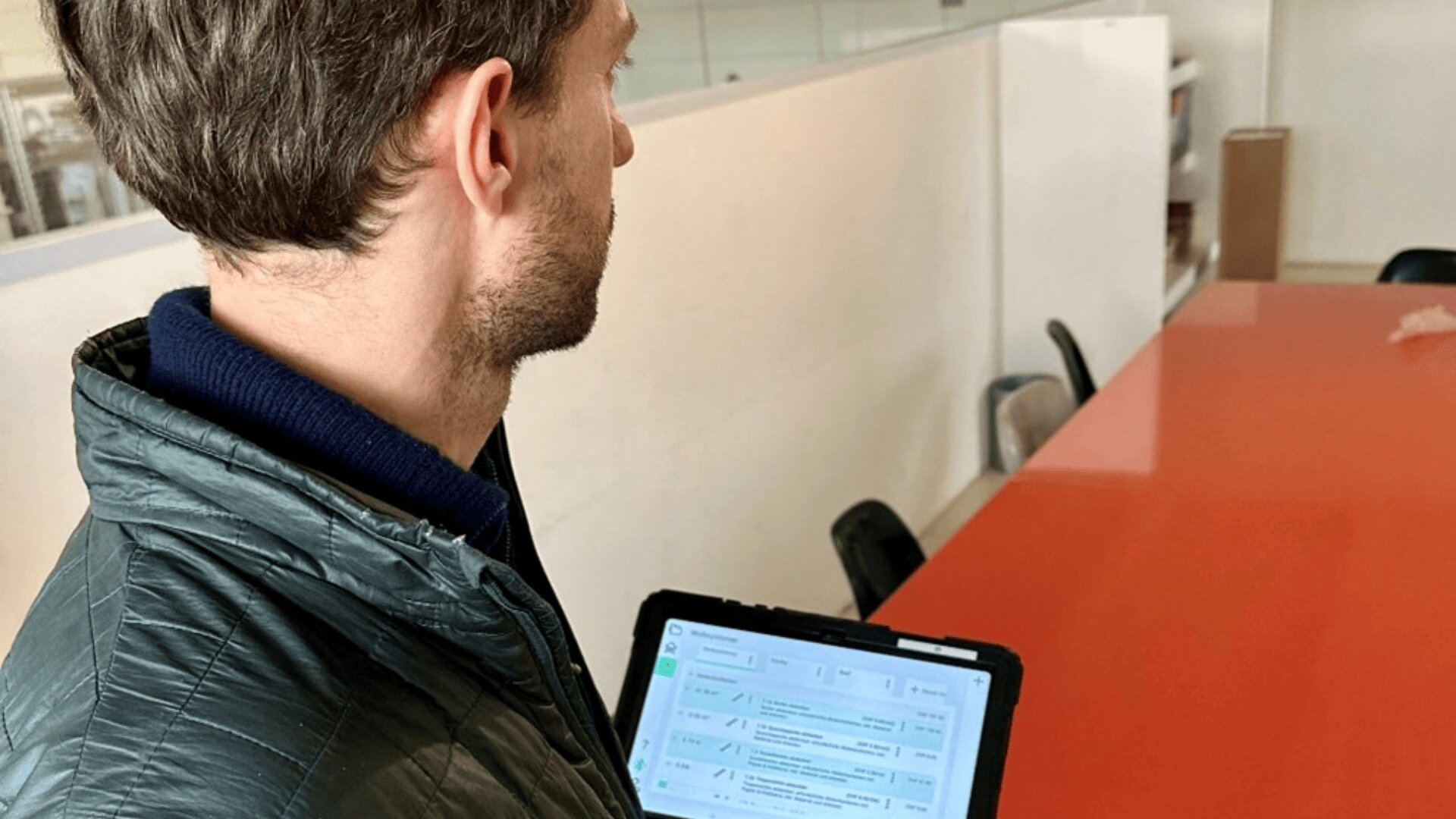 Eine Person mit kurzen braunen Haaren hält ein Tablet mit einem digitalen Dokument in der Hand und steht neben einem langen roten Konferenztisch in einer modernen Büroumgebung.