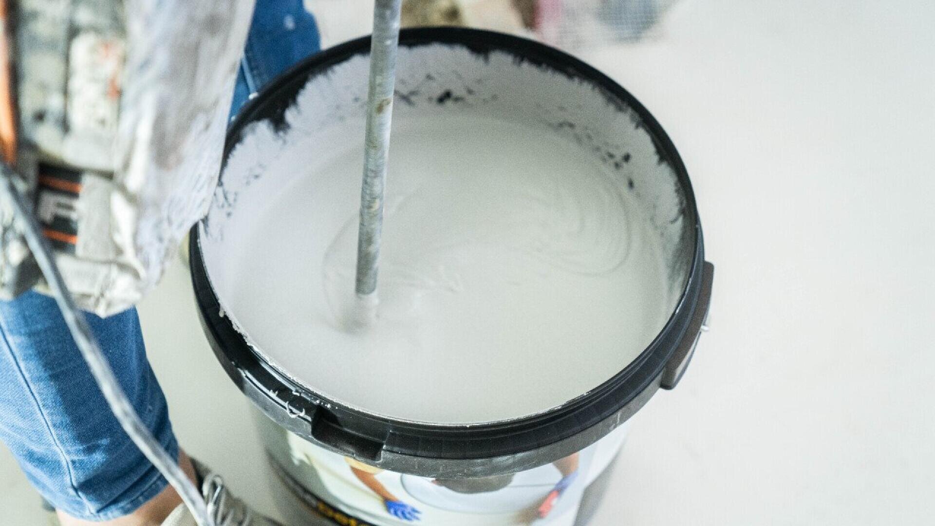 A close-up of a person mixing white paint in a large black bucket using a power mixer. Only part of the persons legs and hands are visible. The scene appears to be part of a painting or renovation project.