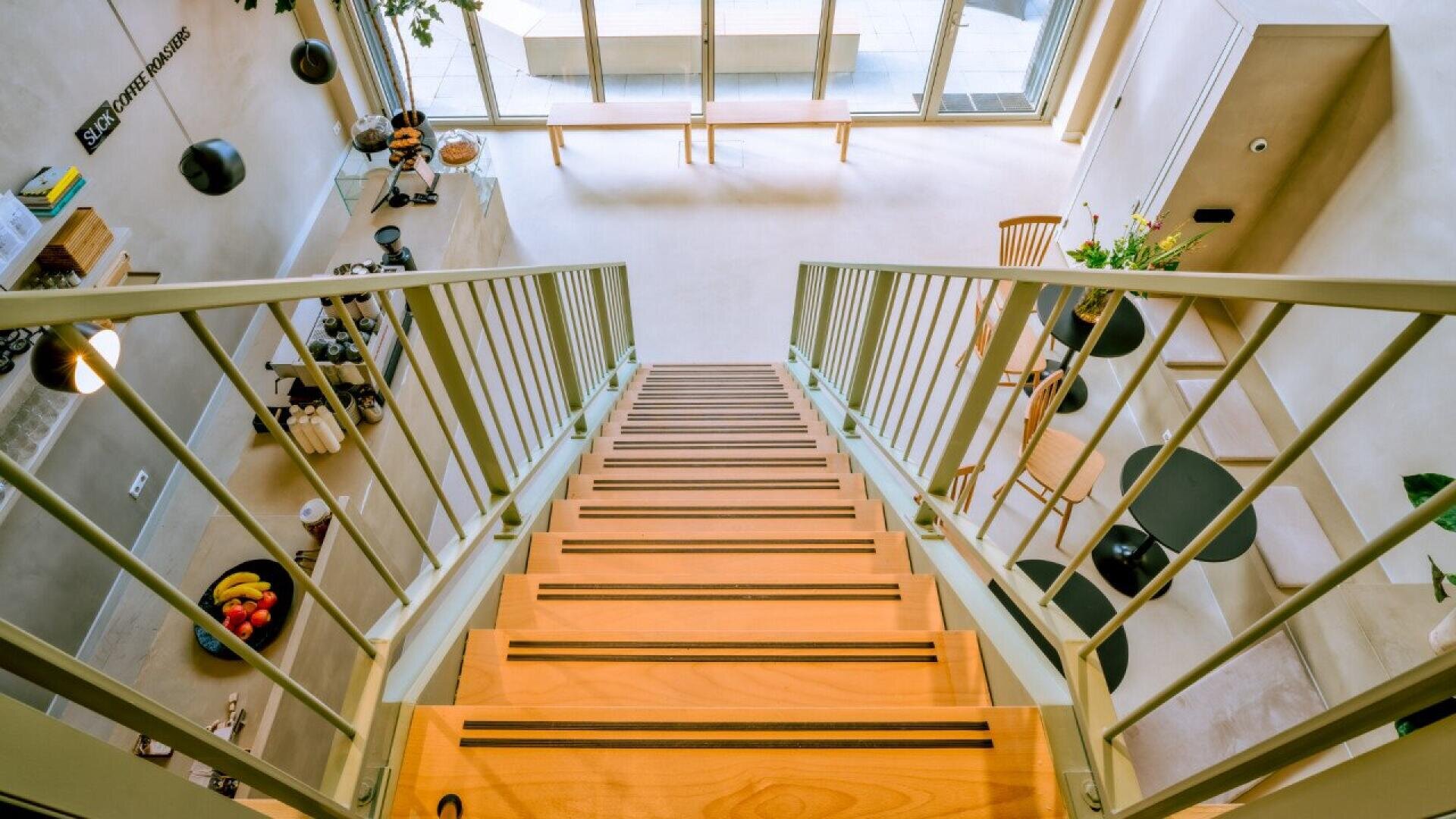 View from the top of a modern indoor staircase with wooden steps and metal railings, overlooking a bright, open living area with large windows, a bench, dining table, chairs, and potted plants.