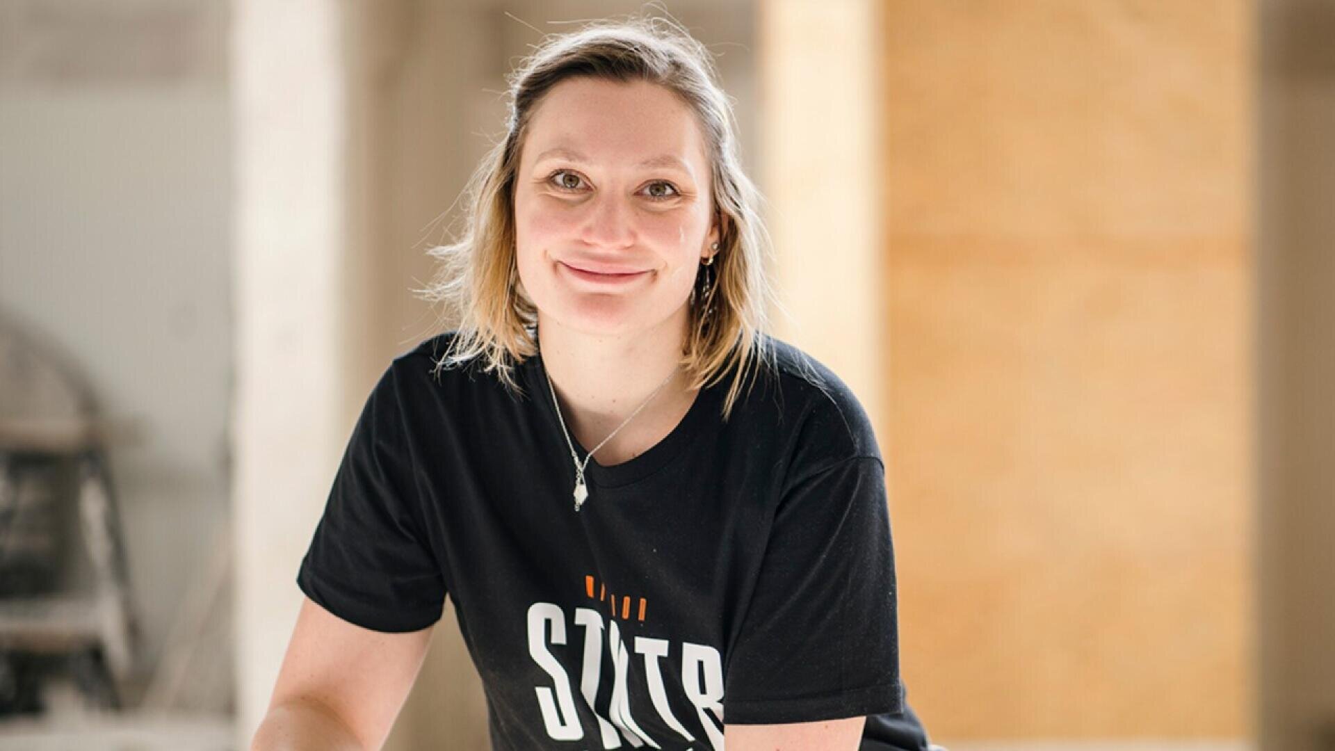 A person with short, light brown hair smiles while wearing a black T-shirt with text and a necklace, standing indoors in a softly lit room with neutral-colored walls.