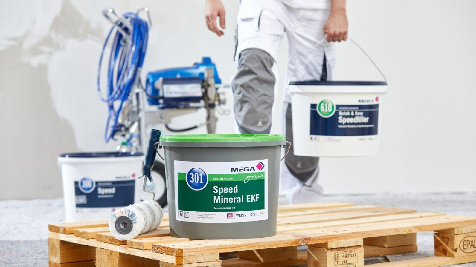 A person in white work pants carries two buckets of paint, with more paint containers and a paint sprayer on a wooden pallet in a bright room under renovation.