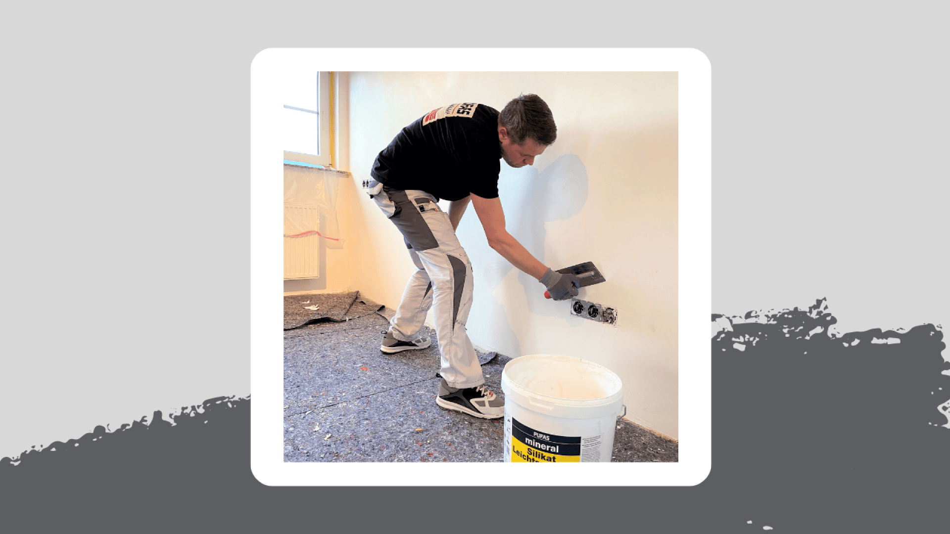Eine Person in Arbeitskleidung trägt mit einer Kelle Putz oder Spachtelmasse auf eine weiße Wand in der Nähe von Steckdosen auf. Ein großer weißer Behälter steht daneben auf dem Boden. Der Raum scheint gerade renoviert zu werden.