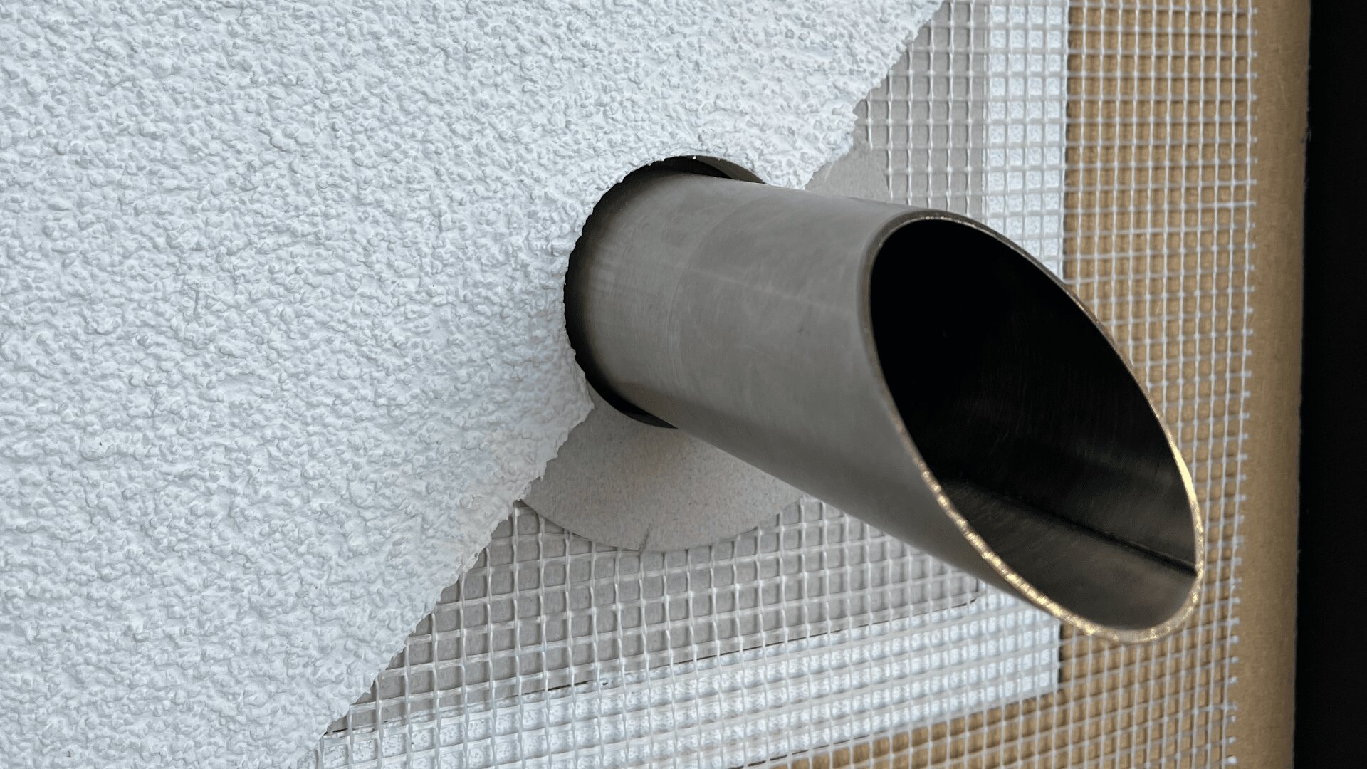 A metal pipe protrudes through a textured white wall with a circular opening. The wall is partially covered by a grid-like mesh material, and the pipe is angled downward.