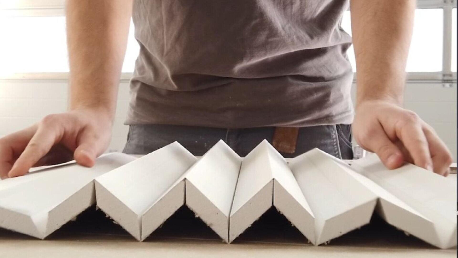 A person in a gray shirt arranges or holds several white, zigzag-shaped pieces of material on a flat surface, creating a geometric, folded pattern.