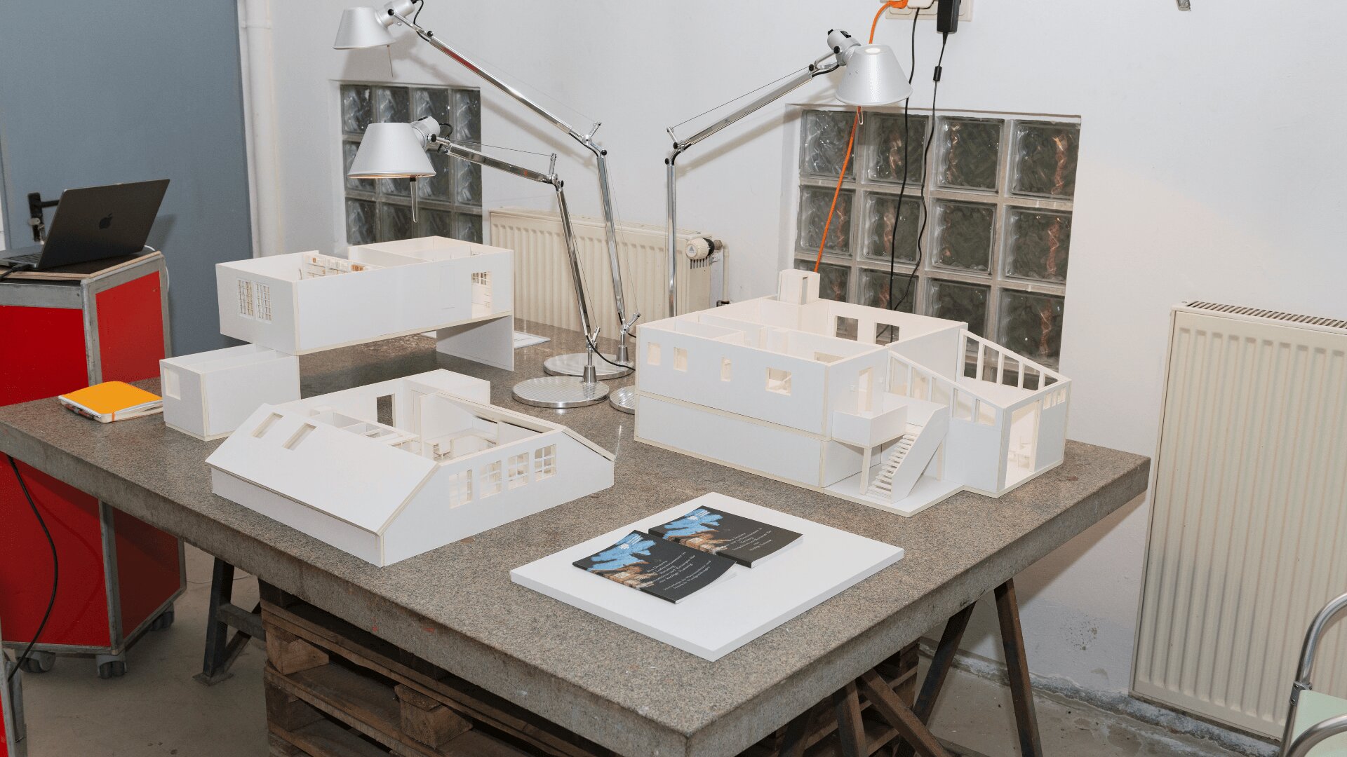 Drei maßstabsgetreue Architekturmodelle moderner Gebäude sind auf einem Arbeitstisch ausgestellt. In der Nähe befinden sich zwei verstellbare Schreibtischlampen, ein Heizkörper und ein roter Schrank. Auf dem Tisch liegt außerdem eine Broschüre oder ein Heft.