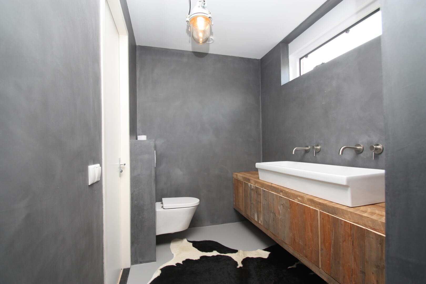 Modernes, minimalistisches Badezimmer mit grauen Wänden, schwebendem Holzwaschtisch, rechteckigem weißen Waschbecken, wandmontierten Wasserhähnen, wandhängender Toilette, kleinem hohen Fenster, Hängelampe und einem schwarz-weißen Kuhfellteppich auf dem Boden.