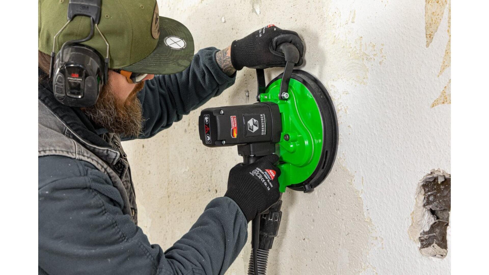 A bearded person wearing safety gear uses a green power tool to sand or grind a white wall, which has some damage and holes.