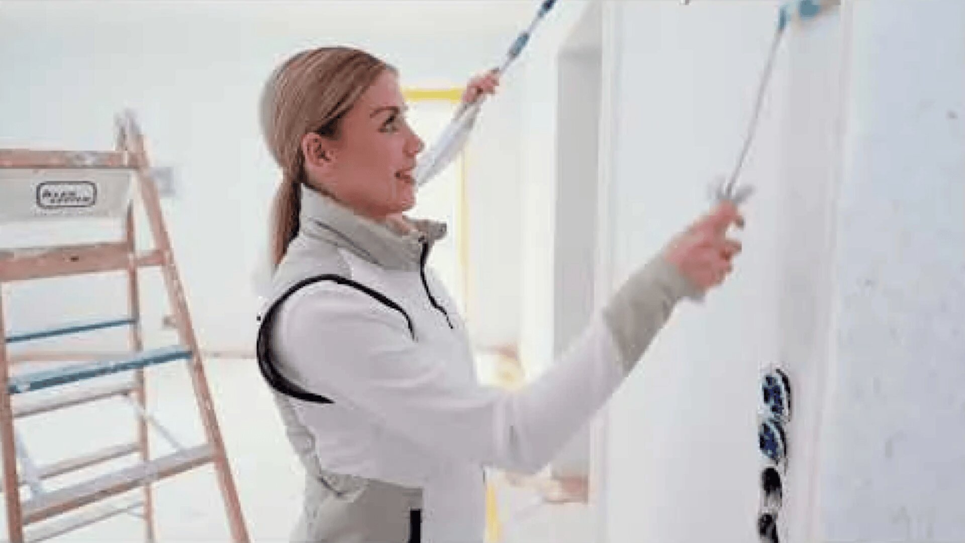 A woman wearing a light-colored outfit paints a white wall with a roller. A ladder is visible in the background in a bright, unfinished room.