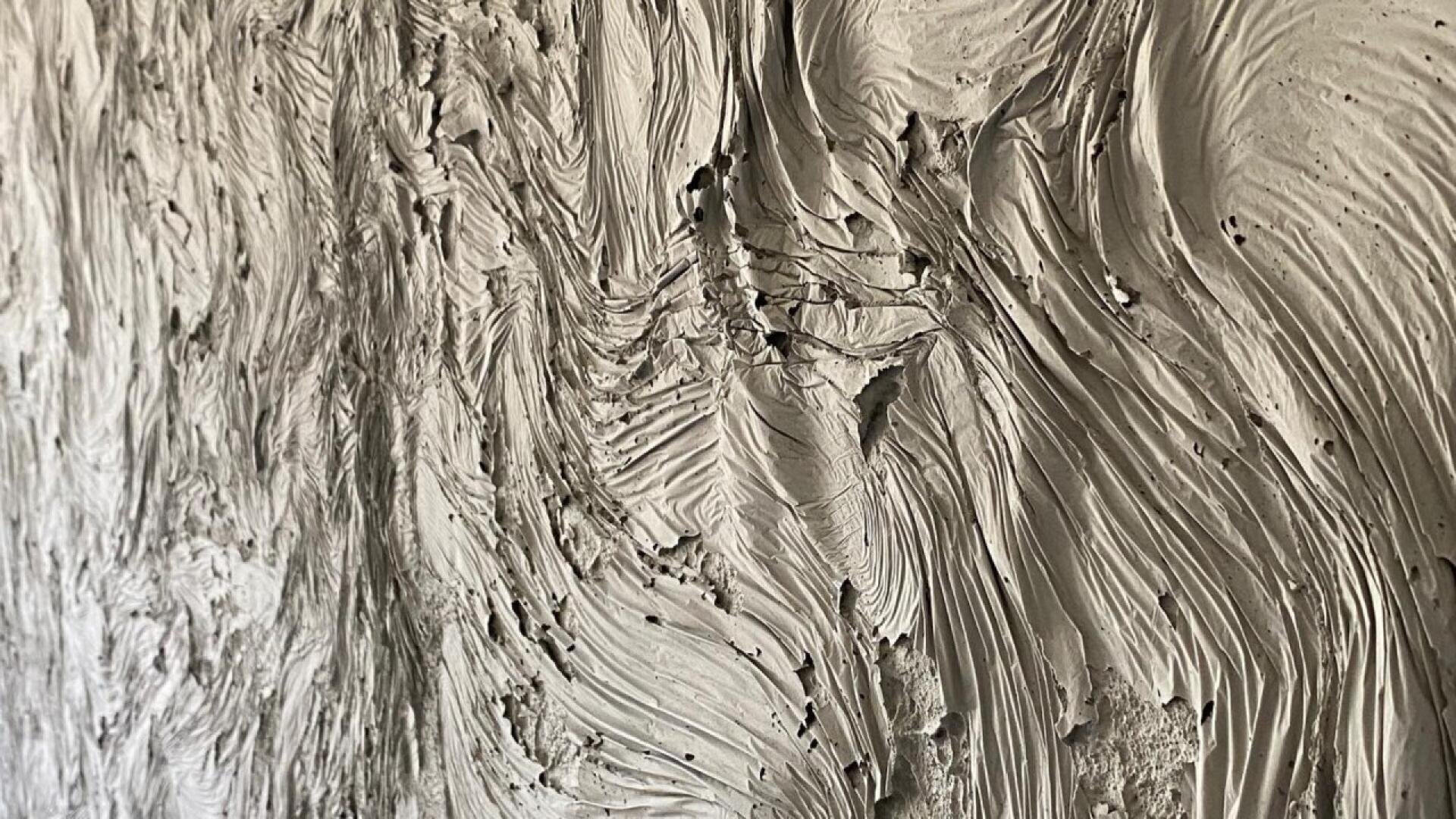 Close-up of textured plaster on a wall, featuring thick, swirling patterns and ridges that create a dynamic, wavy surface. The light grey color highlights the depth and movement of the plaster.