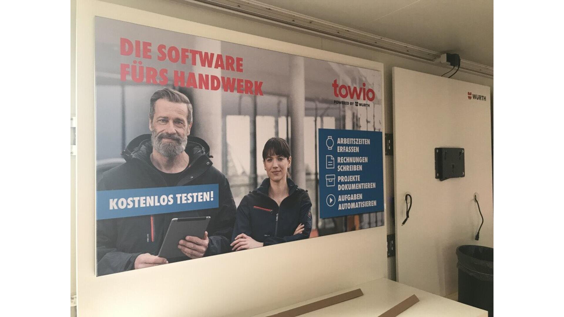 A poster on a wall shows a man and a woman smiling, wearing work jackets. German text promotes software for tradespeople, with features listed and “KOSTENLOS TESTEN!” highlighted. The logo towio is in the top right corner.