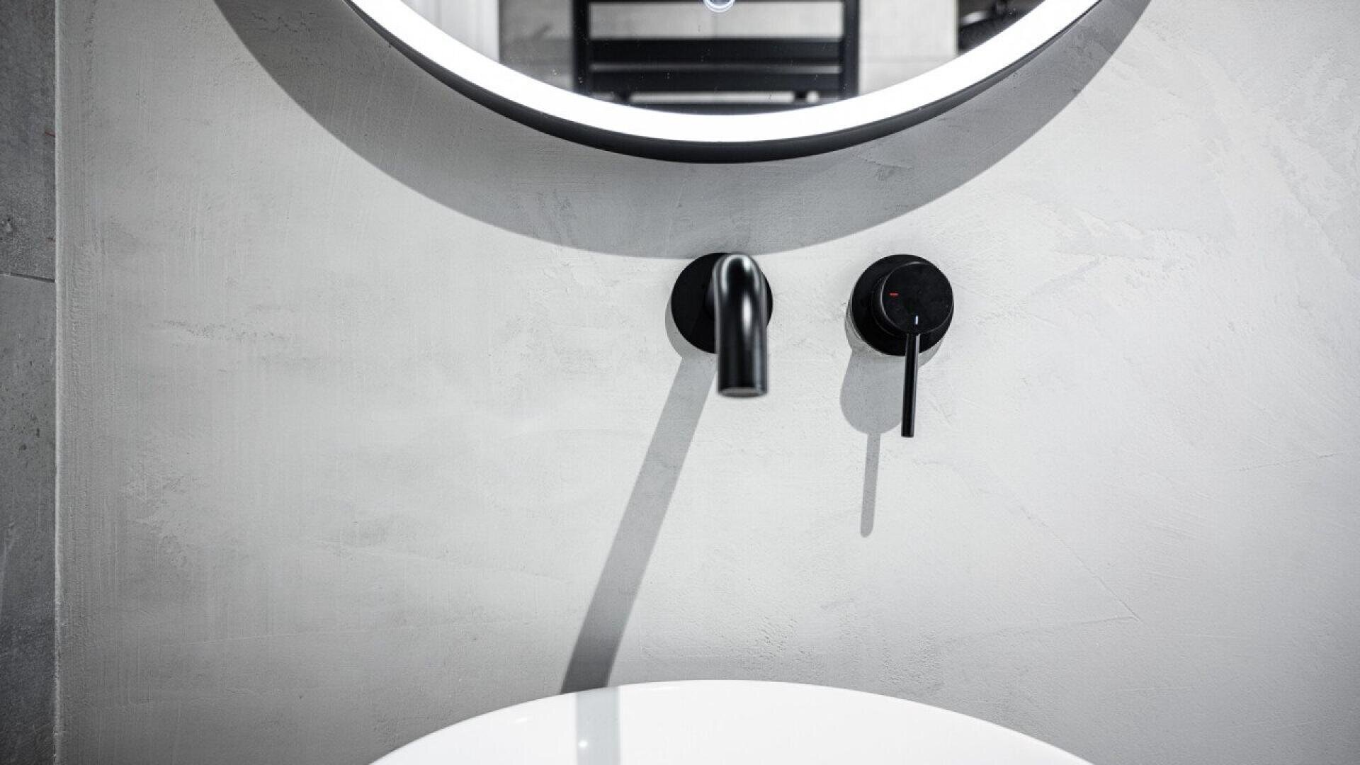 Modern bathroom close-up showing a round mirror with backlighting, a minimalist black wall-mounted faucet, and a matching black handle above a white basin sink against a light gray textured wall.