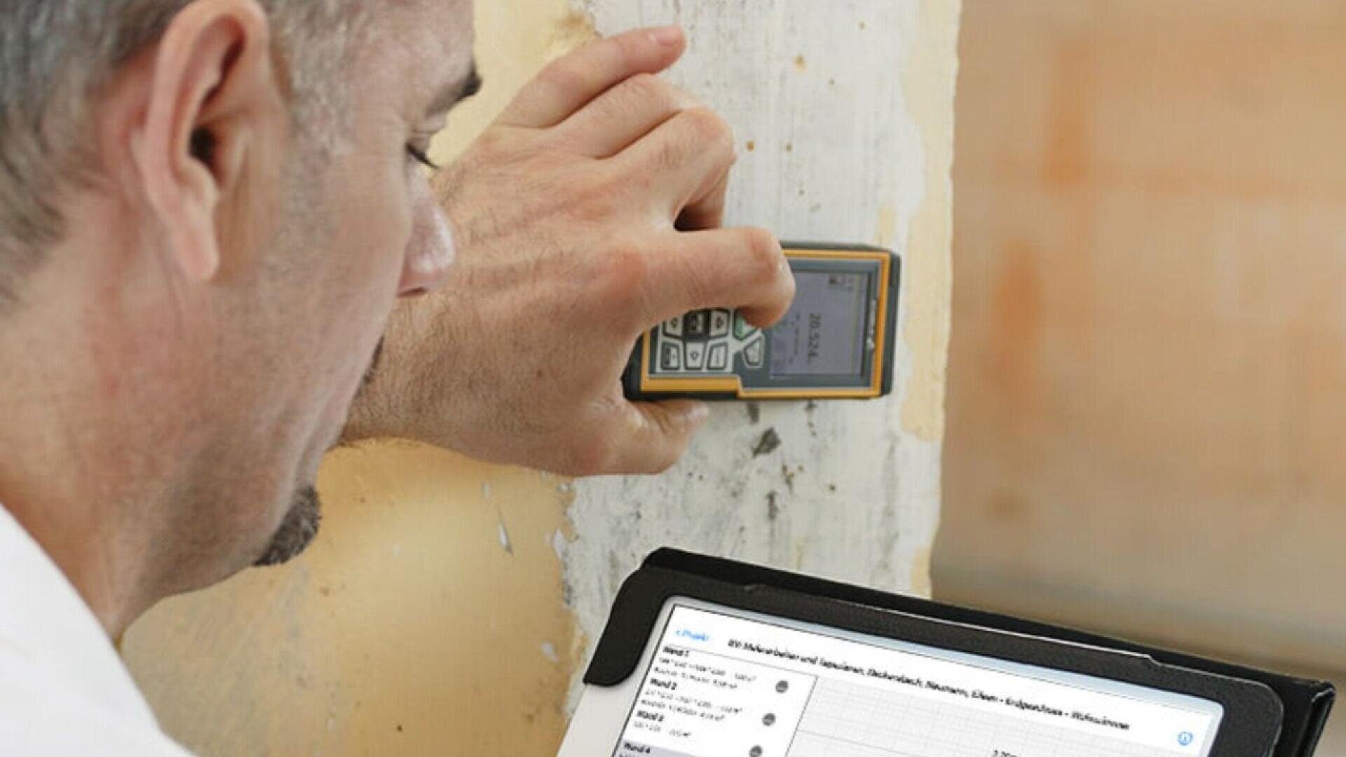 A person uses a handheld measuring device on a wall while viewing data on a tablet, likely recording or analyzing information related to the walls condition.