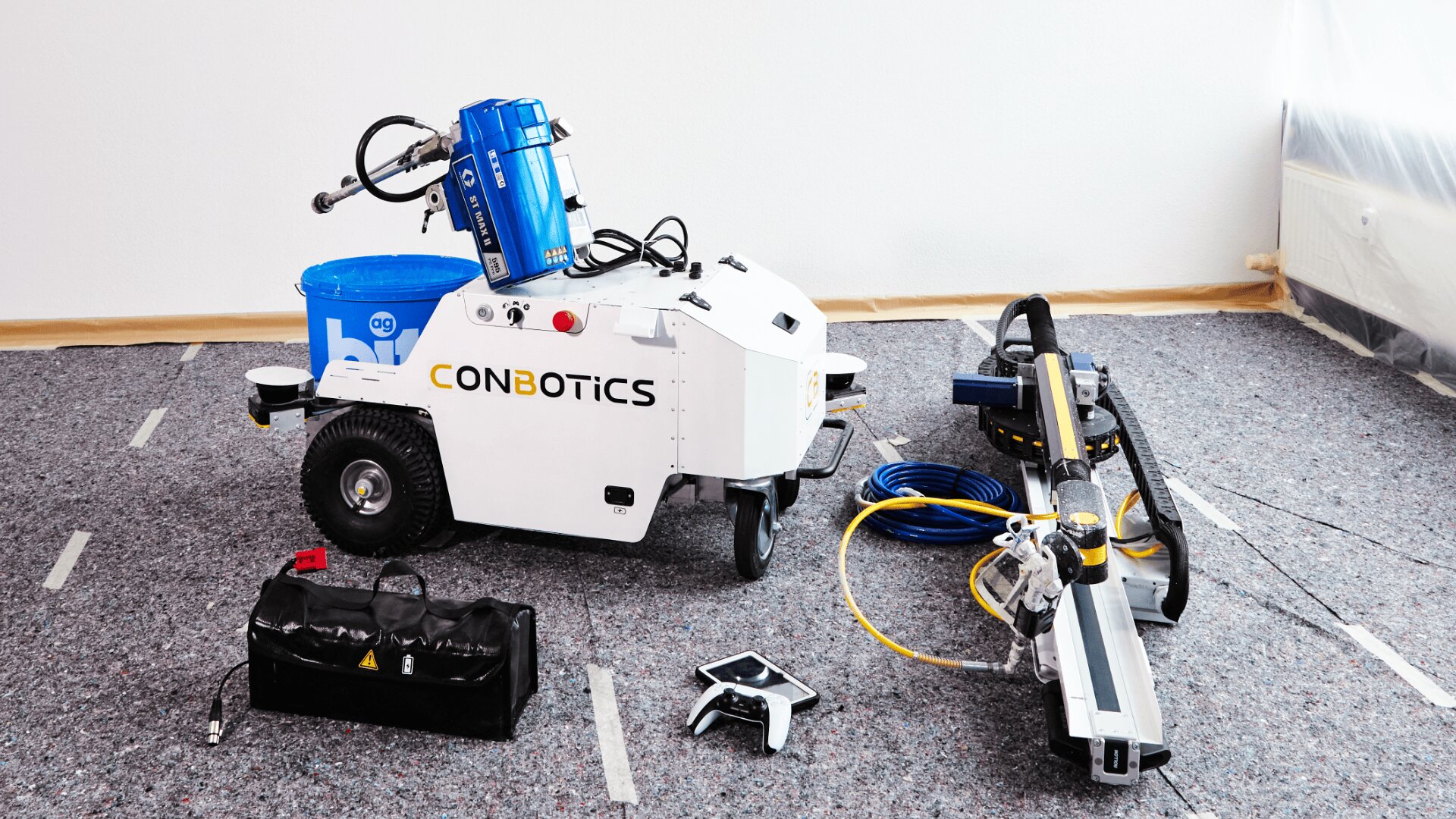 Ein weißer CONBOTICS-Roboter sitzt auf einem Teppichboden neben Werkzeugen, Kabeln, einem blauen Eimer und einem großen grauen Gerät, was auf eine Konstruktion oder einen technischen Aufbau in einer Innenumgebung hindeutet.