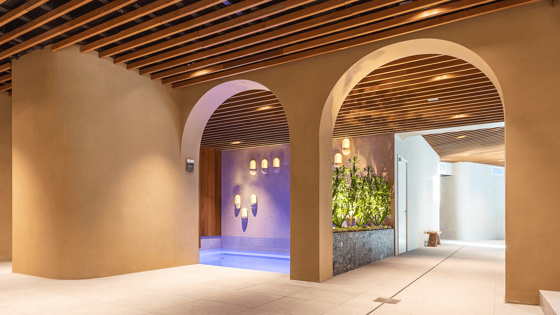 Ein moderner Innenraum mit großen gewölbten Türen, einem kleinen Pool mit Wandleuchten, einem Blumenkasten mit Grünpflanzen und einer Holzlattendecke mit Einbauleuchten.