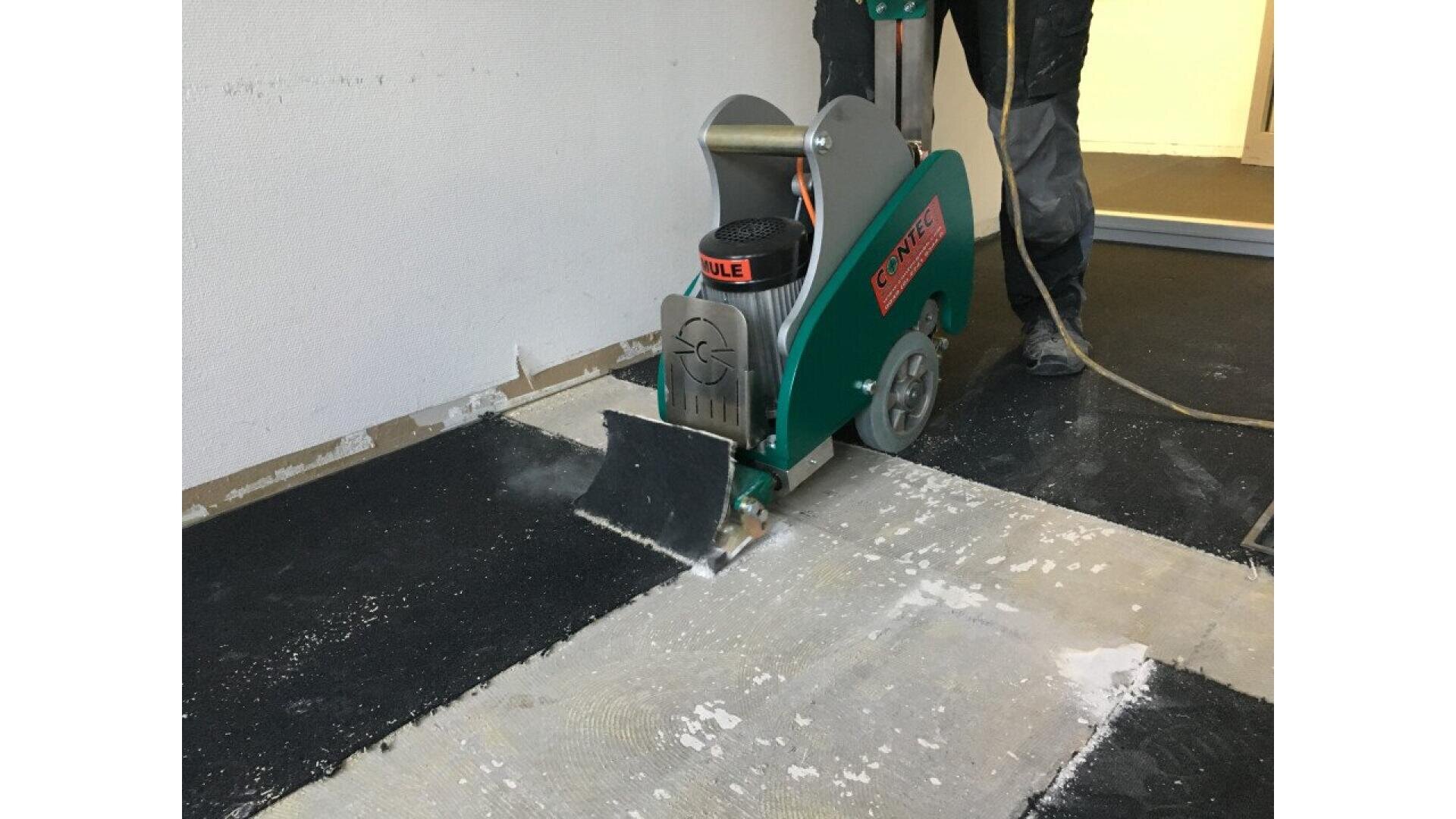 A worker uses a green floor stripping machine to remove old black flooring in a room, with some debris and exposed subfloor visible.