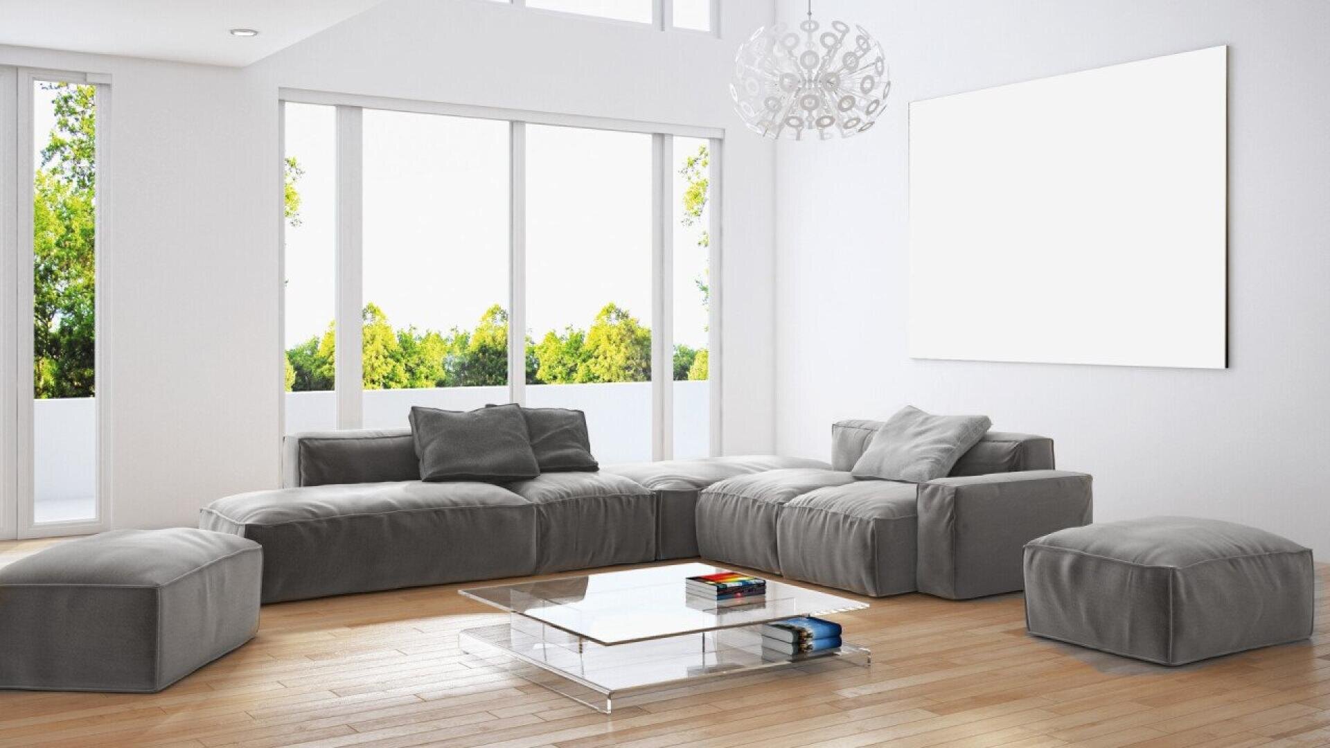 A modern living room with gray sectional sofa, glass coffee table with books, light wood floors, large windows with outdoor greenery, and a blank white wall art piece.