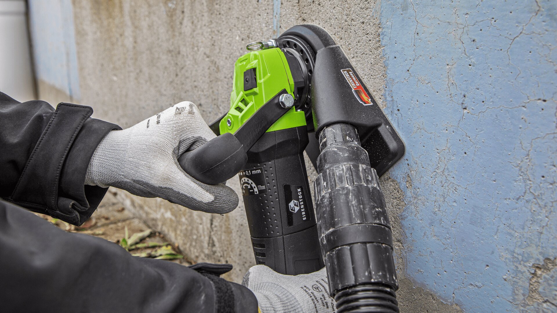 A person wearing protective gloves uses a green and black angle grinder with a dust extraction attachment to sand or grind a concrete wall next to a blue-painted section.