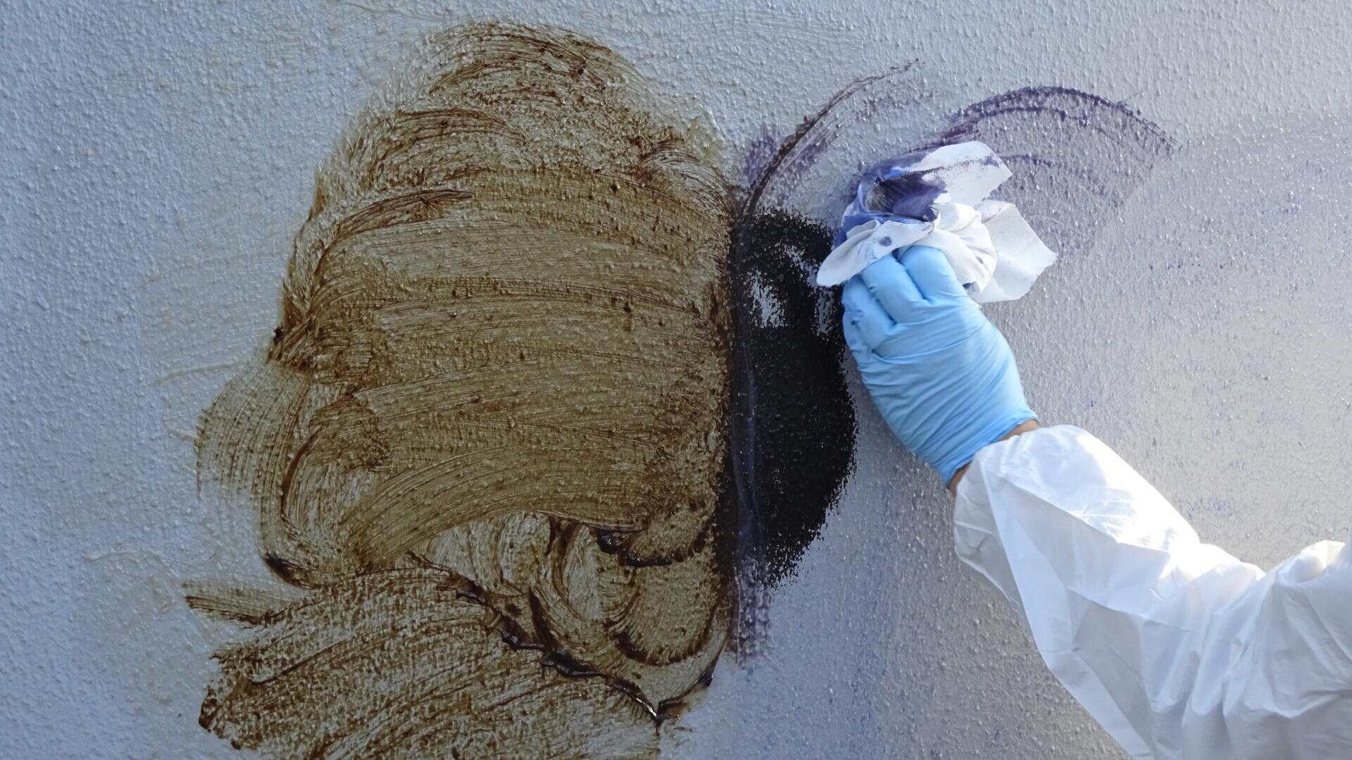 Eine Person mit einem blauen Handschuh und einem weißen Schutzanzug entfernt mit einem Tuch Graffiti von einer strukturierten weißen Wand.