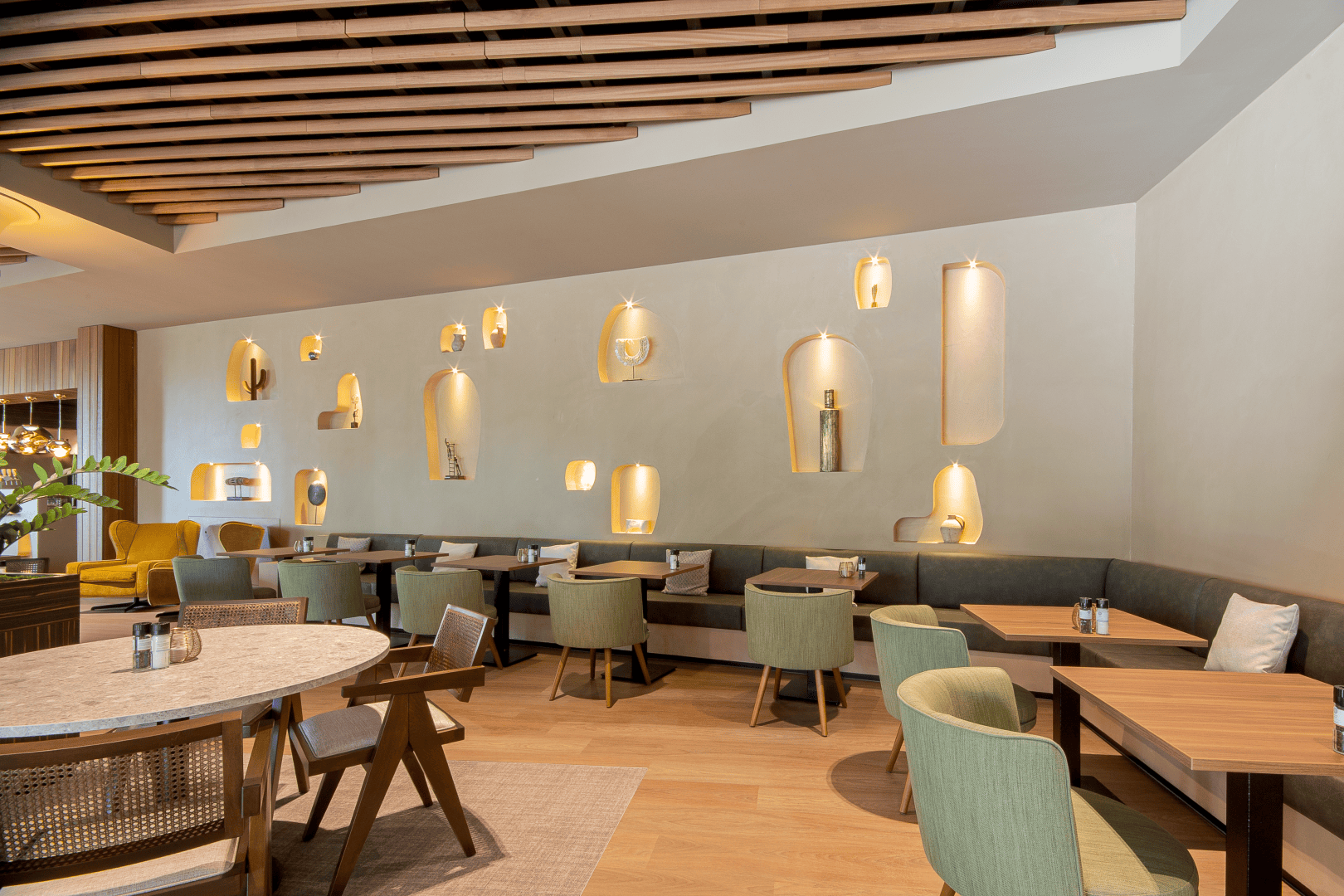 Modernes Restaurant-Interieur mit Holztischen, grünen und hölzernen Stühlen und einer Wand mit dekorativ beleuchteten Nischen, in denen Flaschen und Vasen ausgestellt sind. Die Decke ist mit Holzlamellen versehen, und der Raum wirkt warm und einladend.