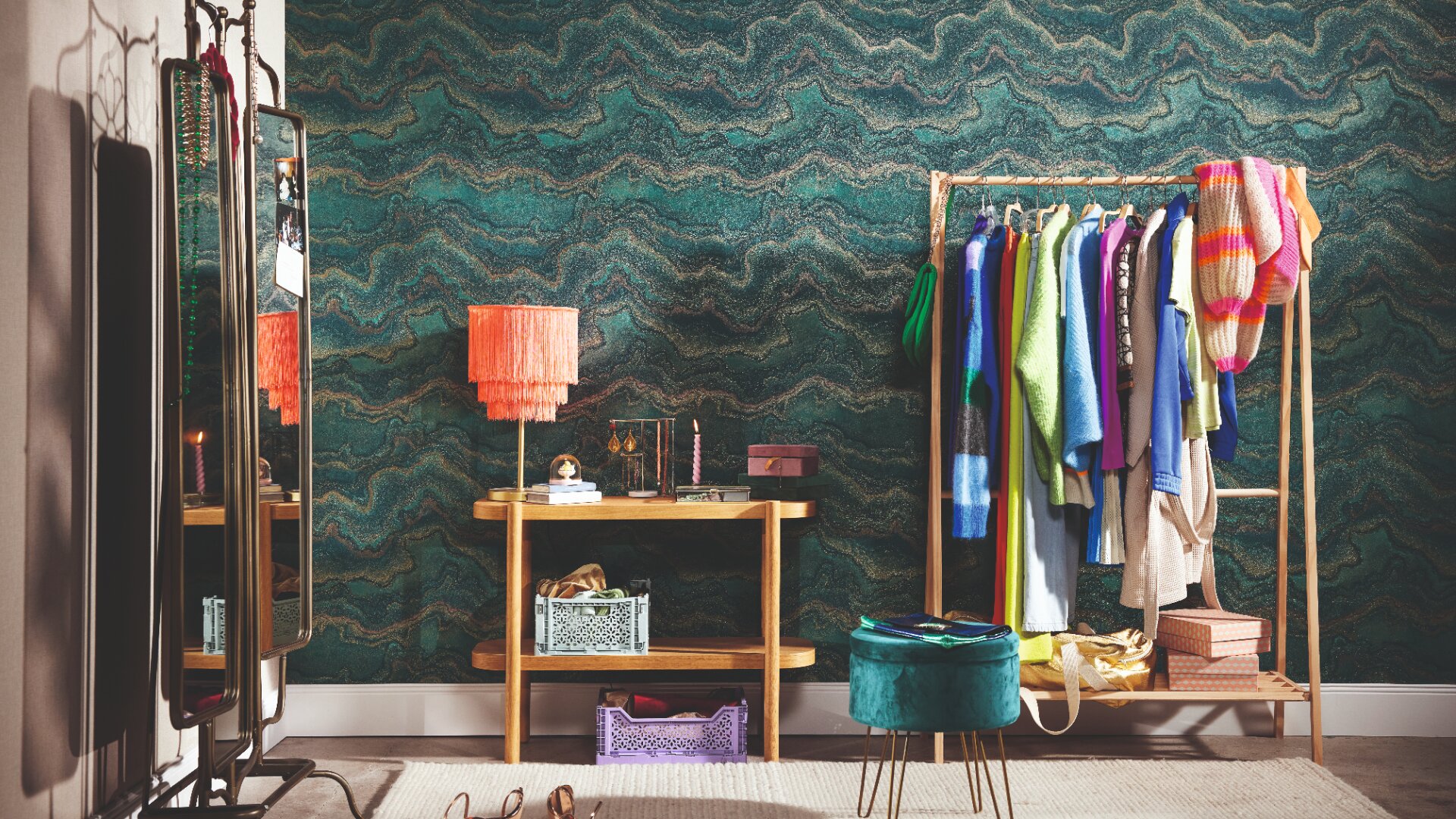 Ein gemütliches Zimmer mit einer gewellten grünen Tapete, einem hölzernen Konsolentisch mit einer orangefarbenen Lampe mit Fransen, einem Standspiegel, einem Kleiderständer mit bunten Kleidern und einem grünen runden Hocker auf einem beigen Teppich.