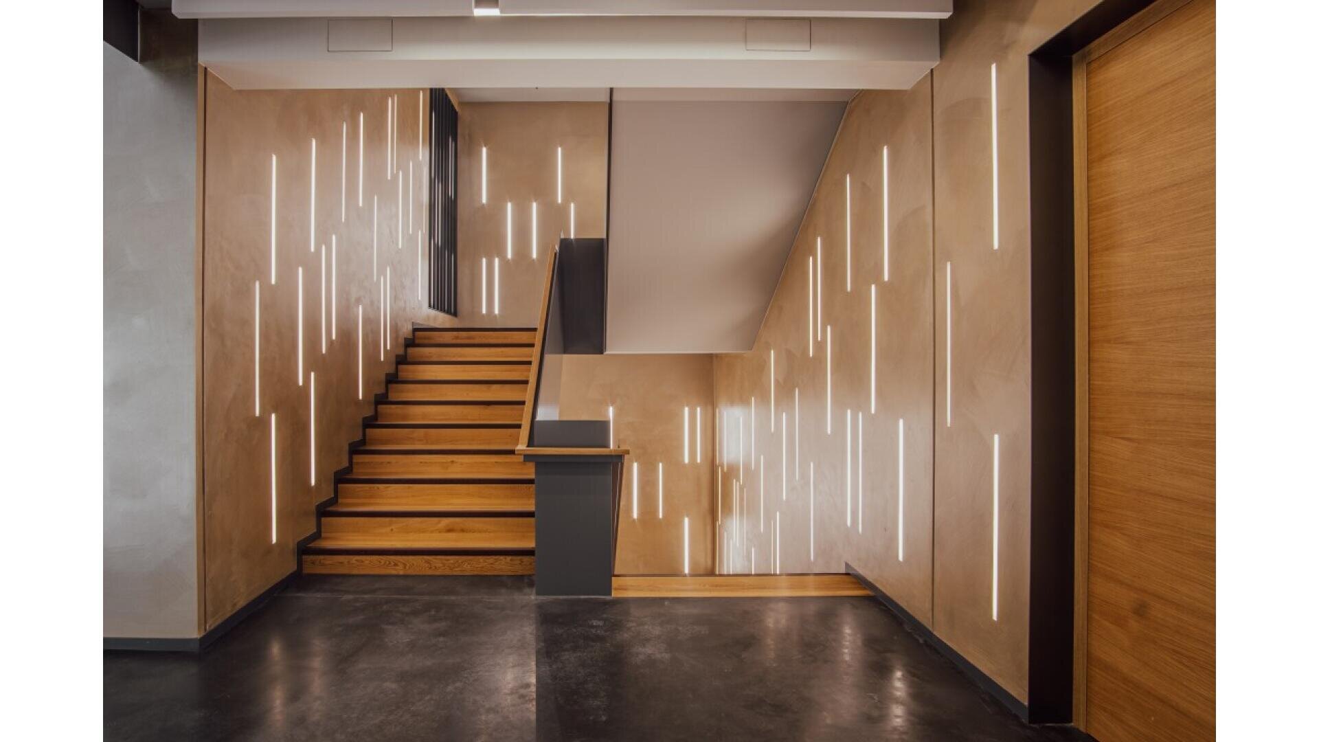 Modern interior with wooden stairs, tan walls featuring vertical recessed LED lights, dark handrails, polished dark floor, and a wooden door on the right. The space has a minimalist, contemporary design.