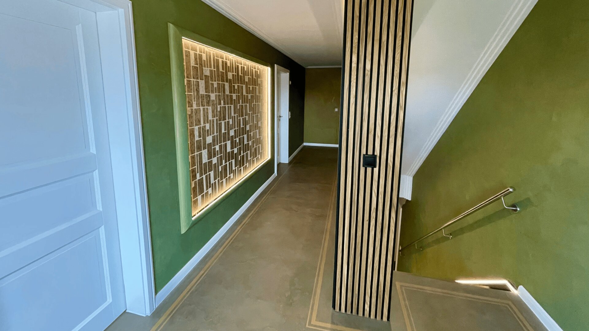 Ein moderner Flur mit grünen Wänden, einer weißen Tür links, einem dekorativen Holzpaneel und einer Treppe mit Metallhandlauf rechts. Der Raum ist gut beleuchtet und besticht durch moderne Designelemente.