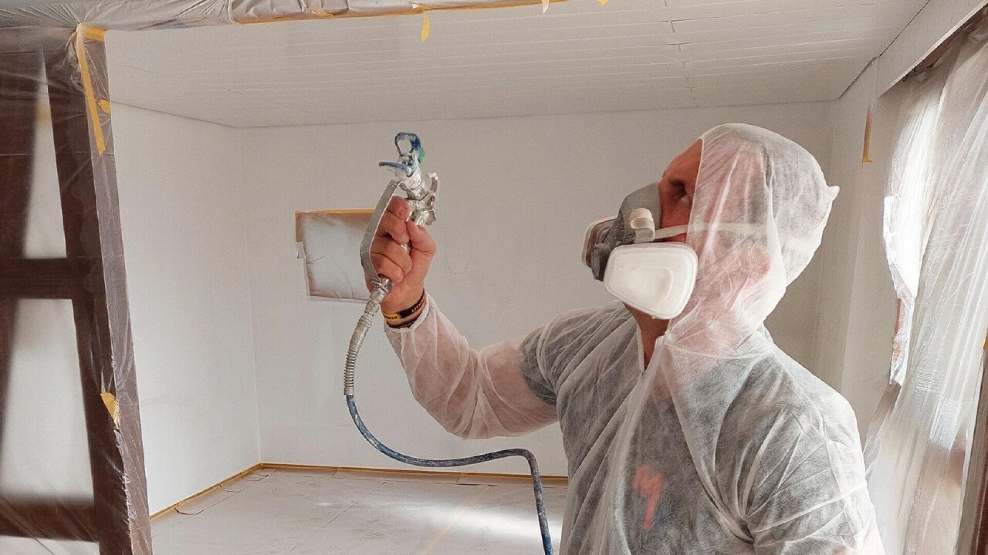 A person wearing protective gear and a respirator mask uses a spray gun to paint the walls of a room covered in plastic sheeting for protection.