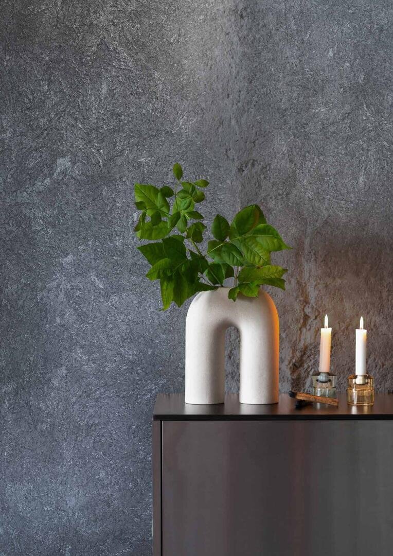 A modern white arch-shaped vase with green leafy branches sits on a dark cabinet beside two lit candles in glass holders against a textured gray wall.