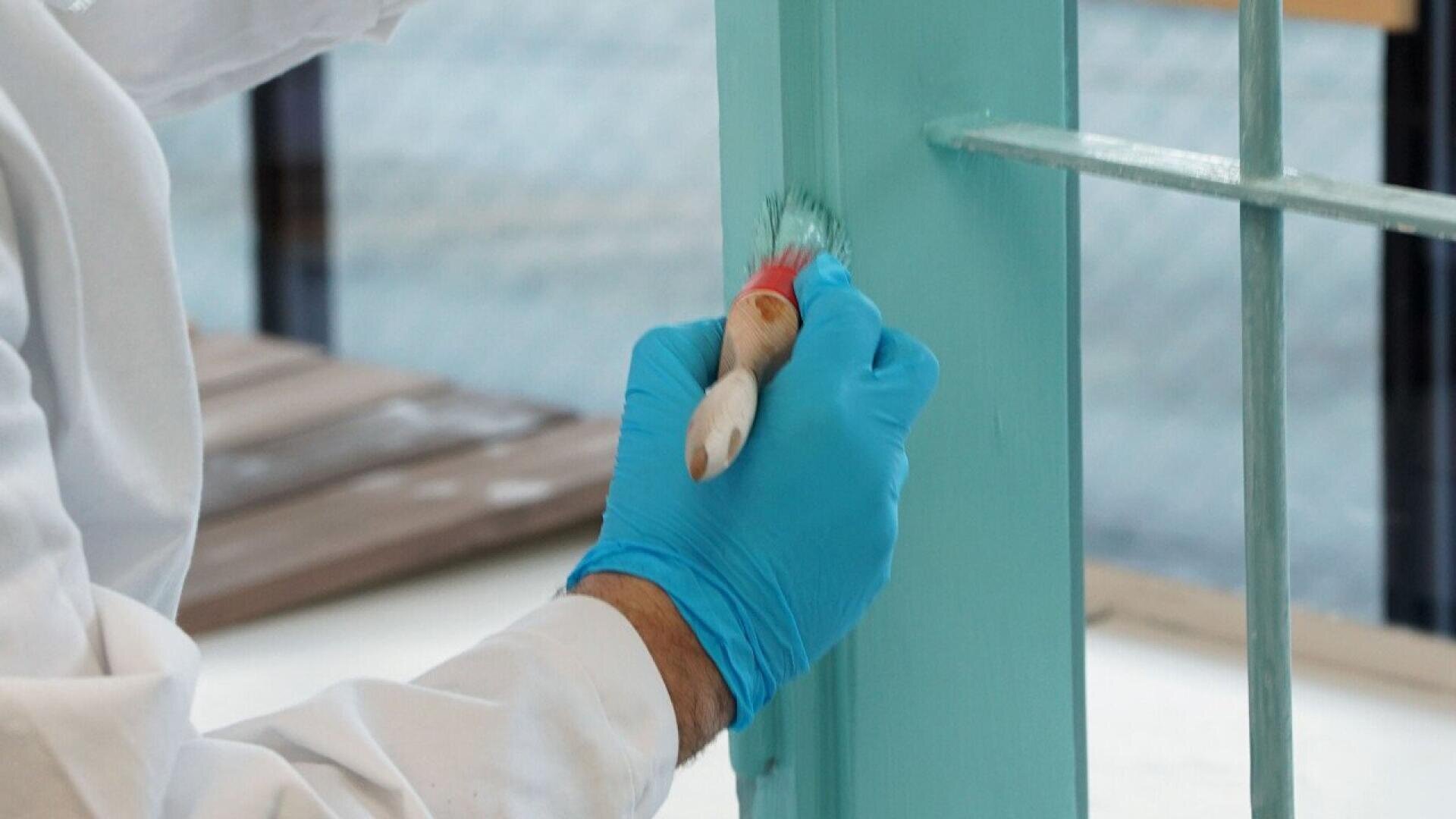 A person wearing blue gloves paints a wooden window frame light turquoise using a small paintbrush.