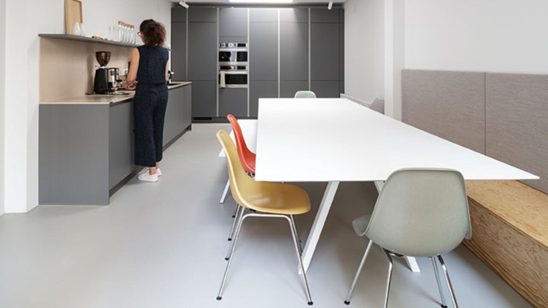 A person stands at a modern kitchen counter preparing coffee. In the foreground is a white dining table with colorful chairs and a built-in wooden bench along one side. The kitchen has grey cabinets and appliances.