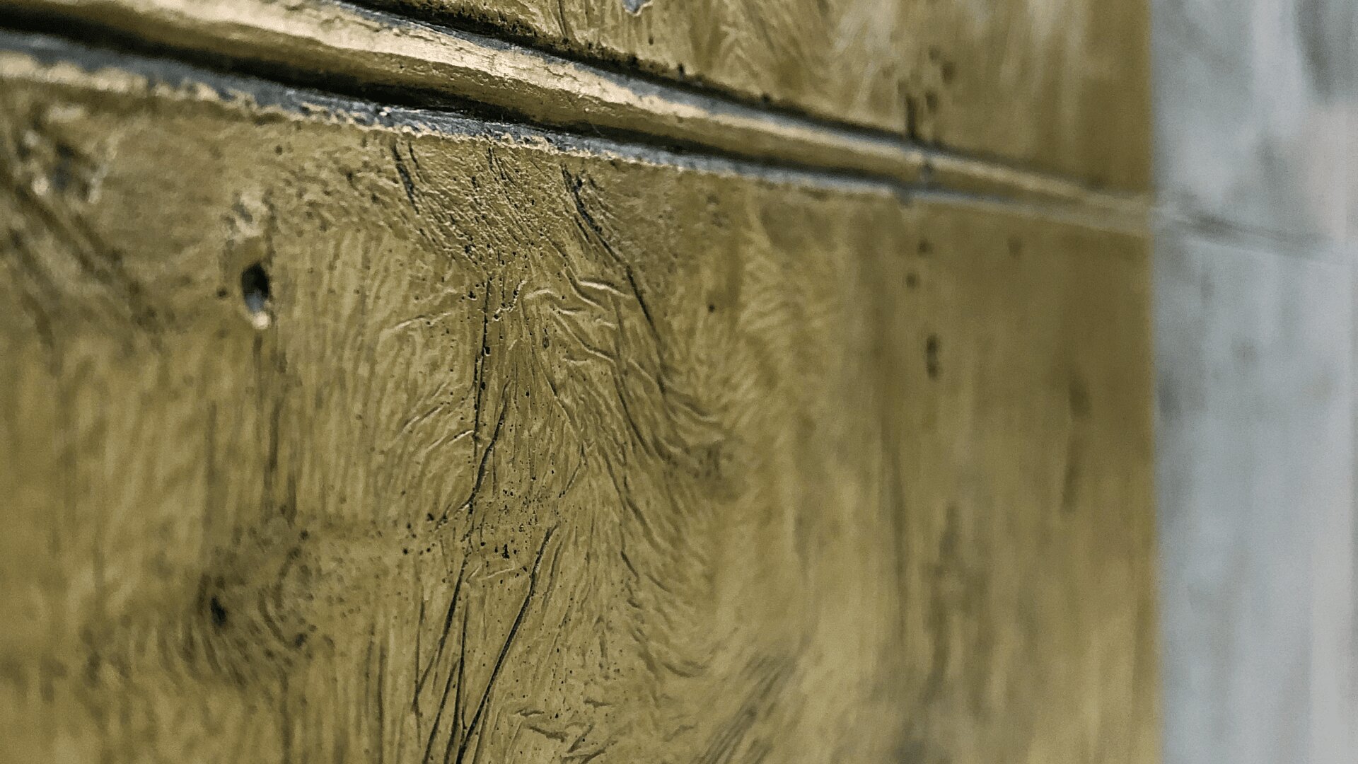 Nahaufnahme einer strukturierten Wand mit Goldoberfläche, die Pinselstriche und Rillen zeigt. Eine horizontale Linie teilt die Wand, wobei die linke Seite in Gold und die rechte Seite in einem helleren, gräulichen Farbton gehalten ist.